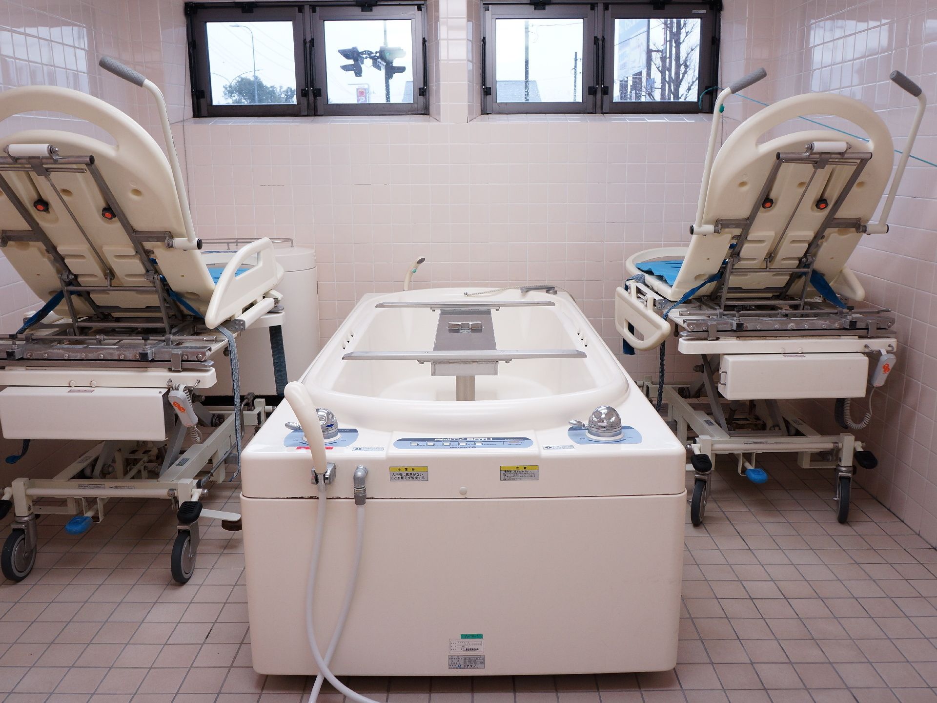 介助用個室バスがある。お風呂で使うリフターが2台両脇に置いてある。床、壁がタイル張りである。窓が4つある。