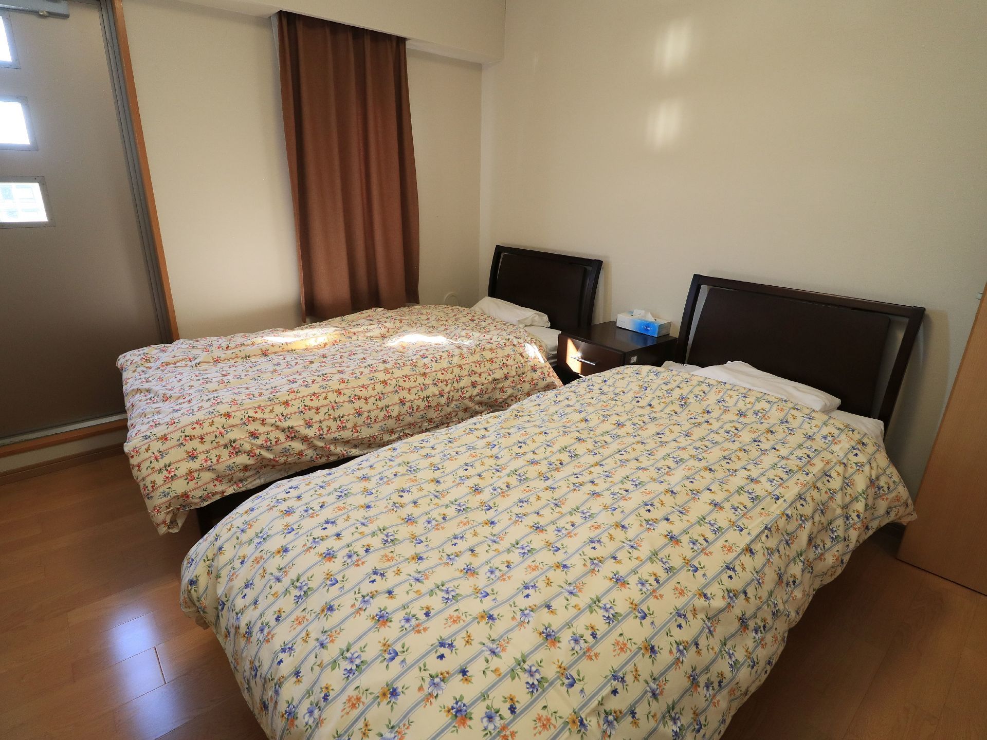 ベッドが２台完備された２人部屋タイプの居室である。色違いの花柄カバーの掛け布団がセットされている。床はフローリングになっている。