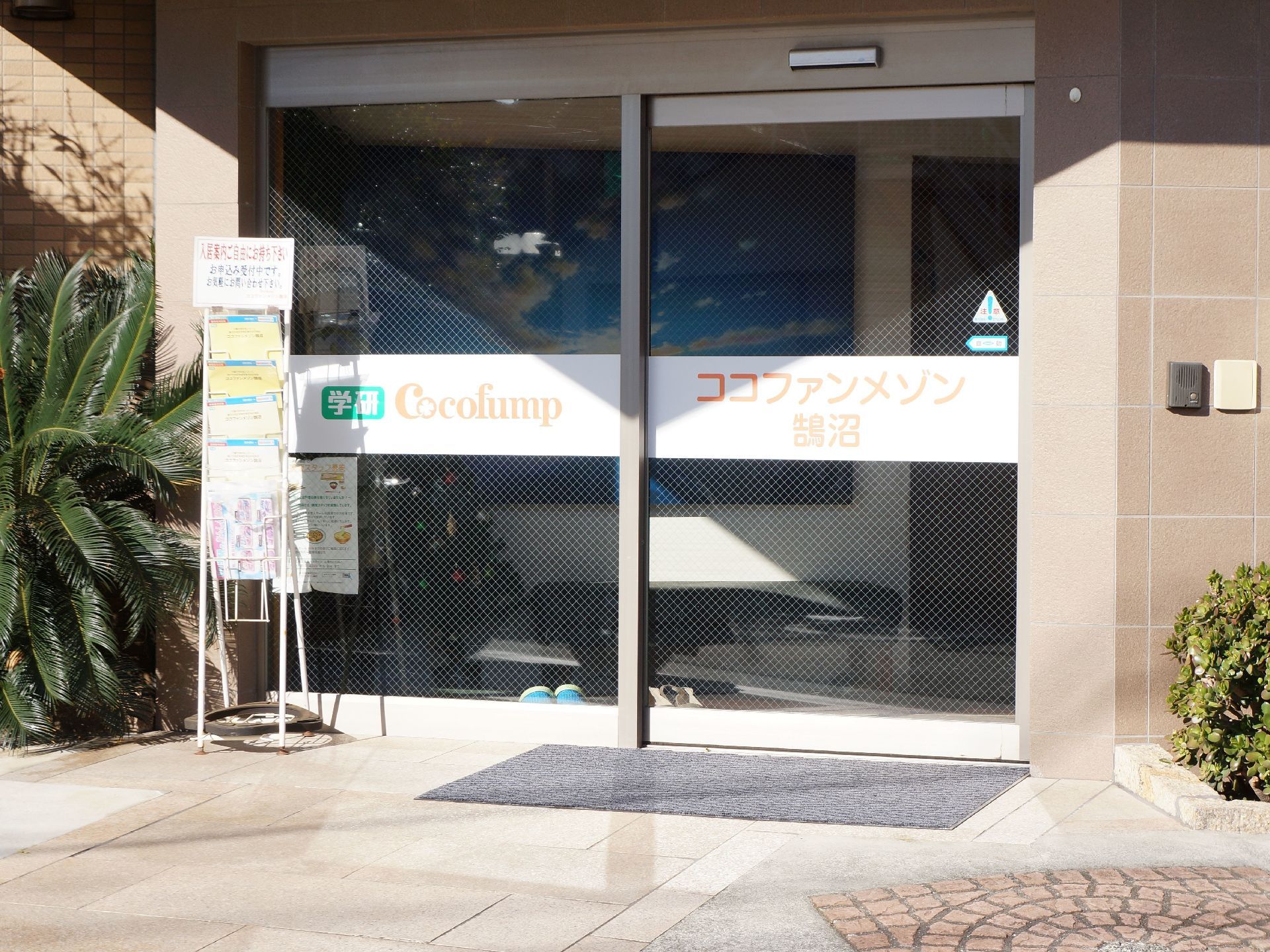 施設の玄関はガラス張りの自動ドアである。前面の床はフラットなタイル敷きで、左側にはパンフレット差しを置いている。