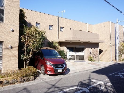 ブラウン系の外壁の建物が２階建ての建物で敷地内には駐車場があり、車が止まっている。