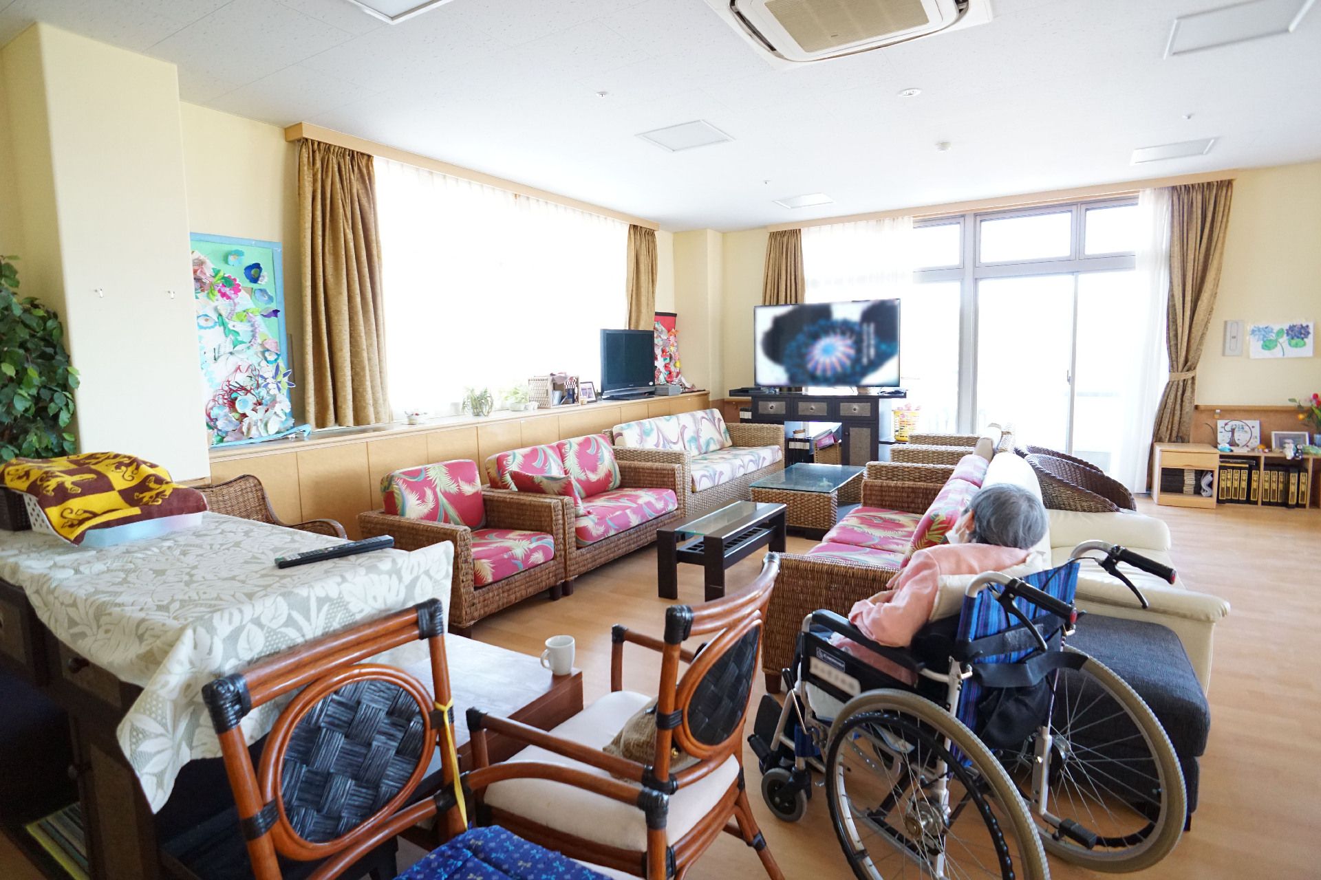 壁２面が窓ガラスに囲まれた広間に車いすで座っている入居者様