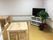 木工で作られたようなシンプルなテーブルや椅子の置かれた部屋。部屋の隅にはテレビ台に乗ったテレビと、土に板が挿してある観葉植物がある。