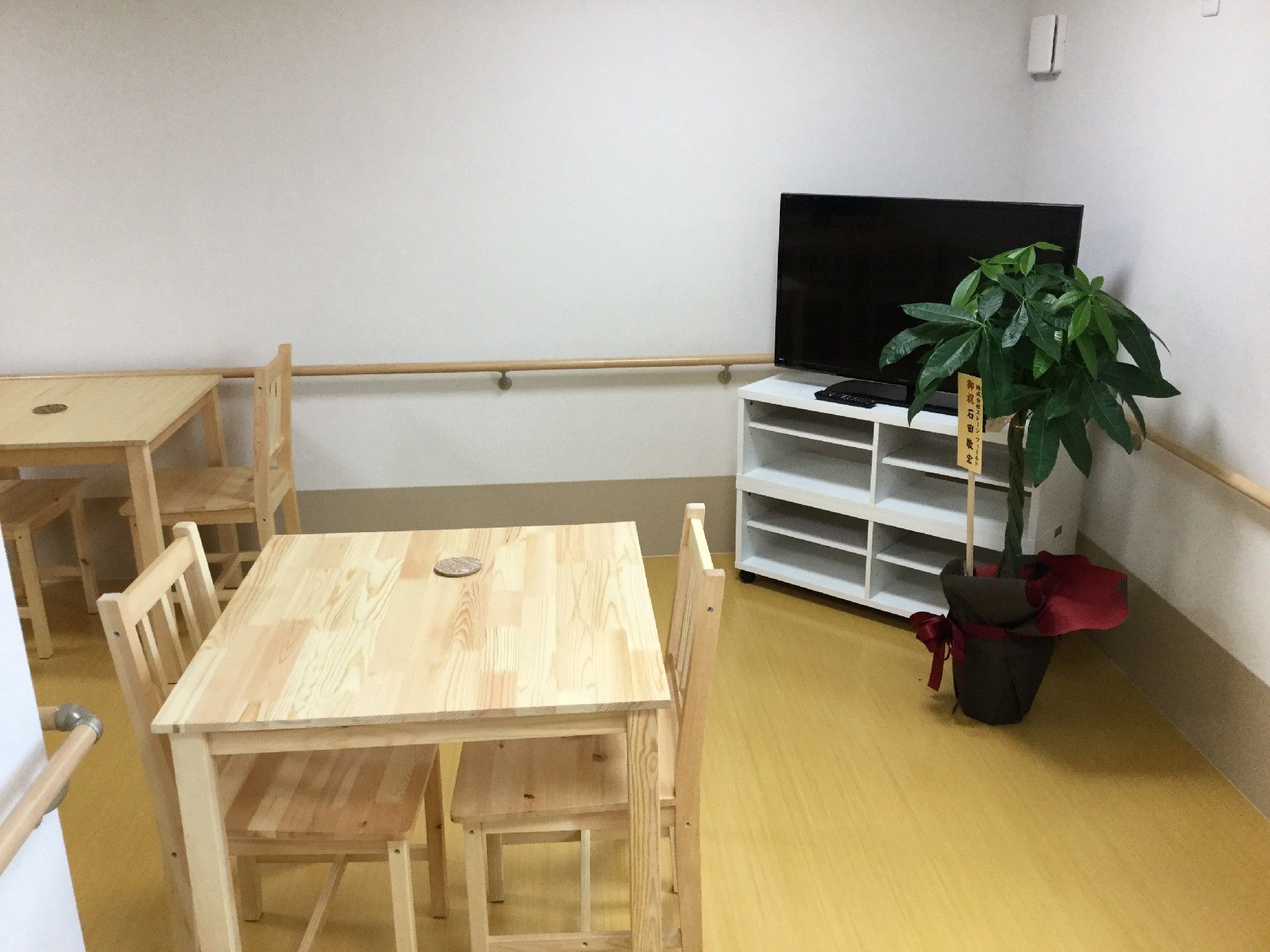 白い壁のコーナーにテレビが設置され、木製のテーブルセットが置かれた共有スペース