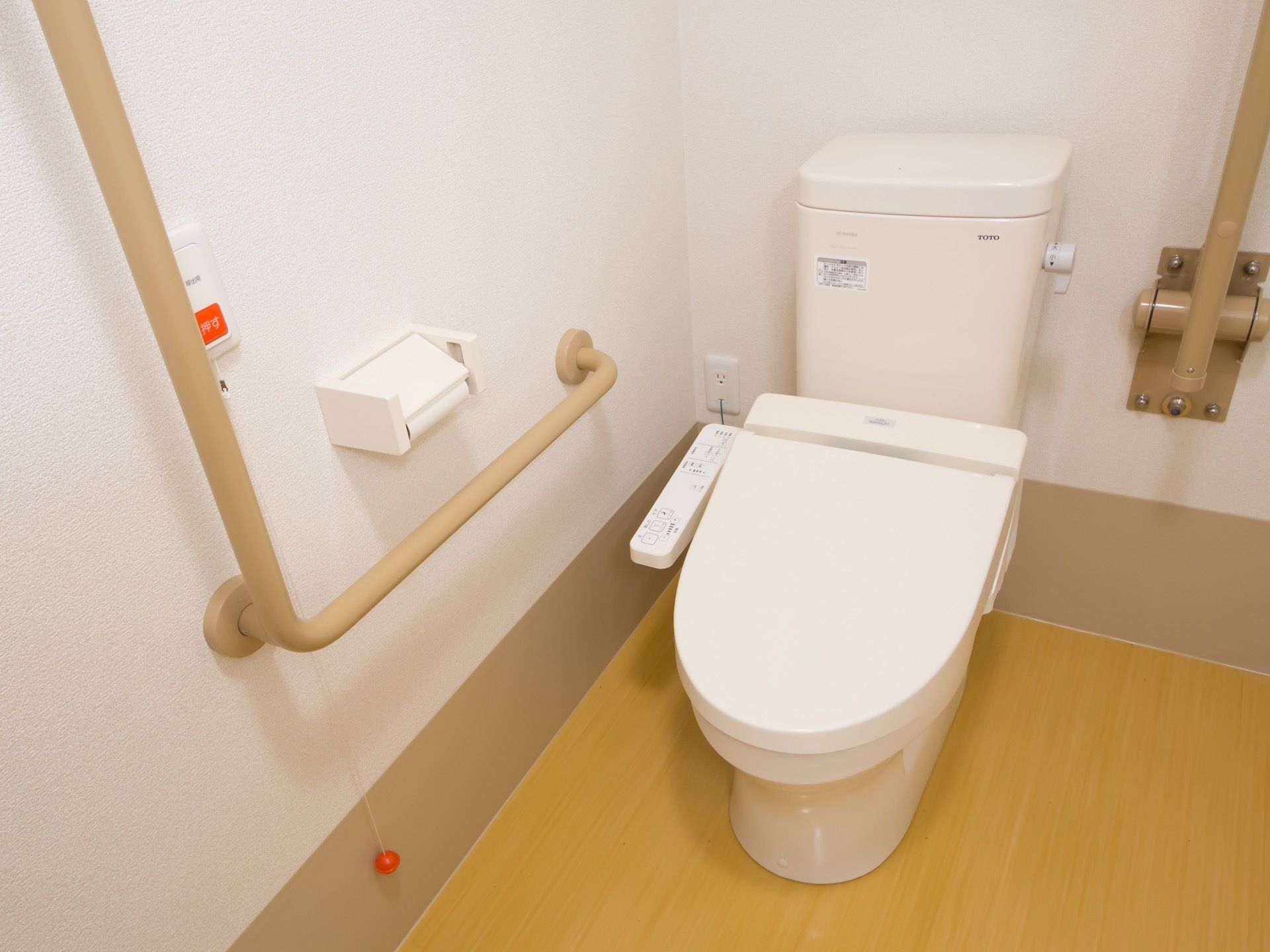 洋式トイレの脇の壁に、L字型の手すりとナースコールが備わっている。もう片側には可動式の手すりが設置されている。