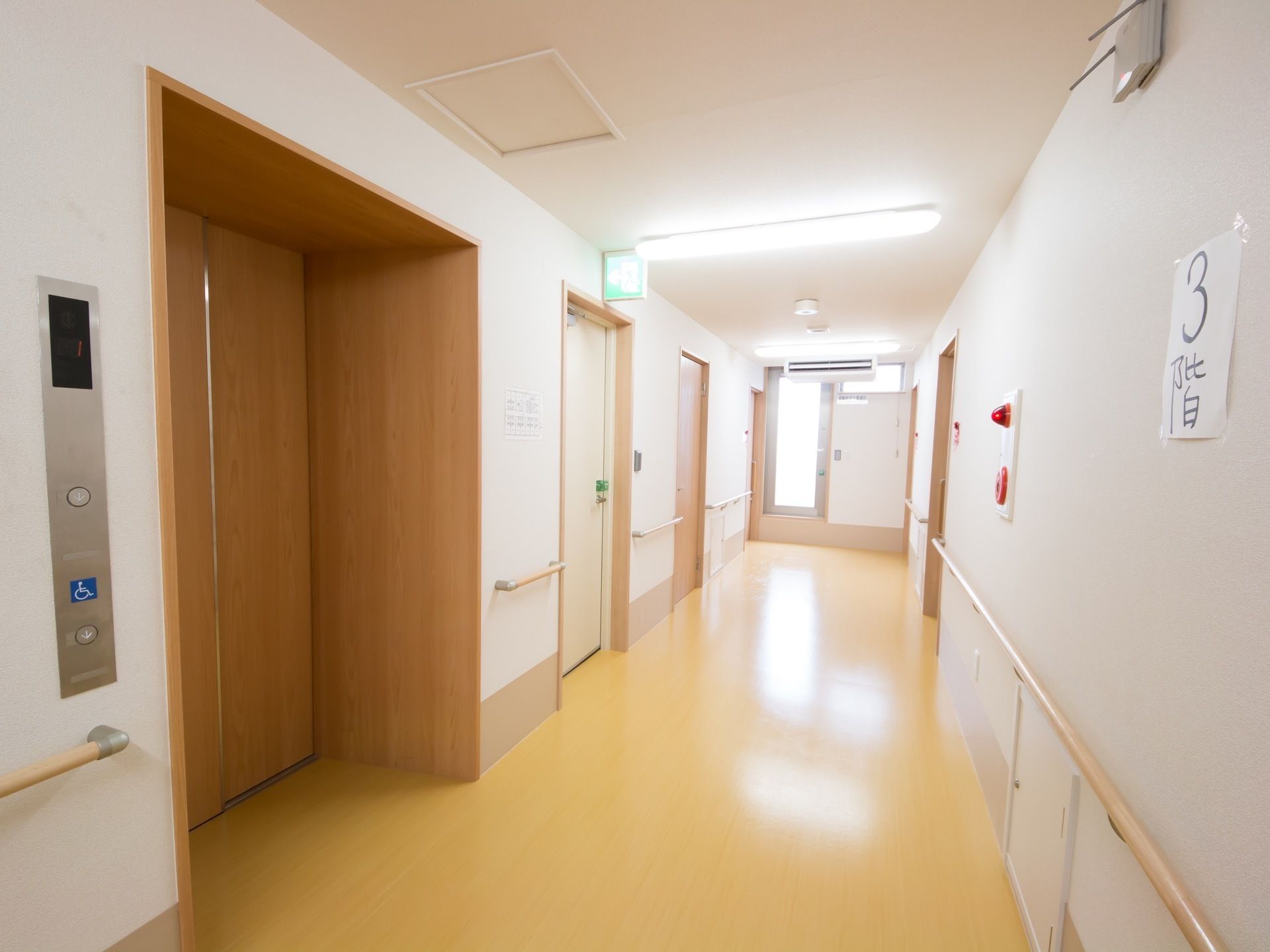 廊下にはエアコンがあり、両側の壁に手すりが設置されている。左側にエレベーターがあり、正面の壁に階数の表示がされている。