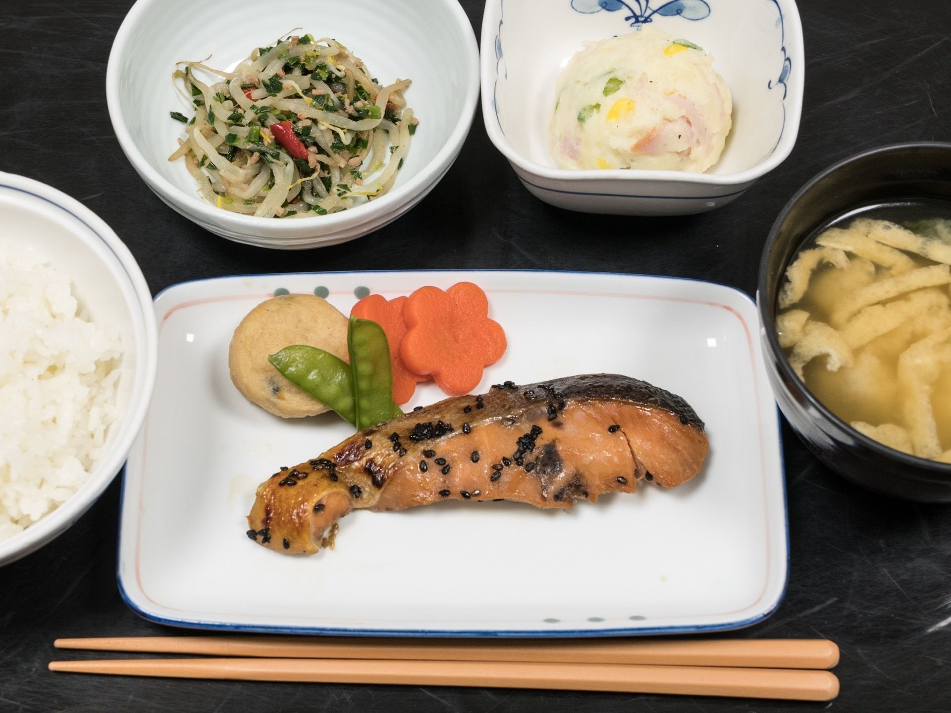 黒いお盆の上に、食事が載っている。メインの鮭に、ポテトサラダや煮物、野菜など、たくさんの食材を使った彩も美しいメニューである。