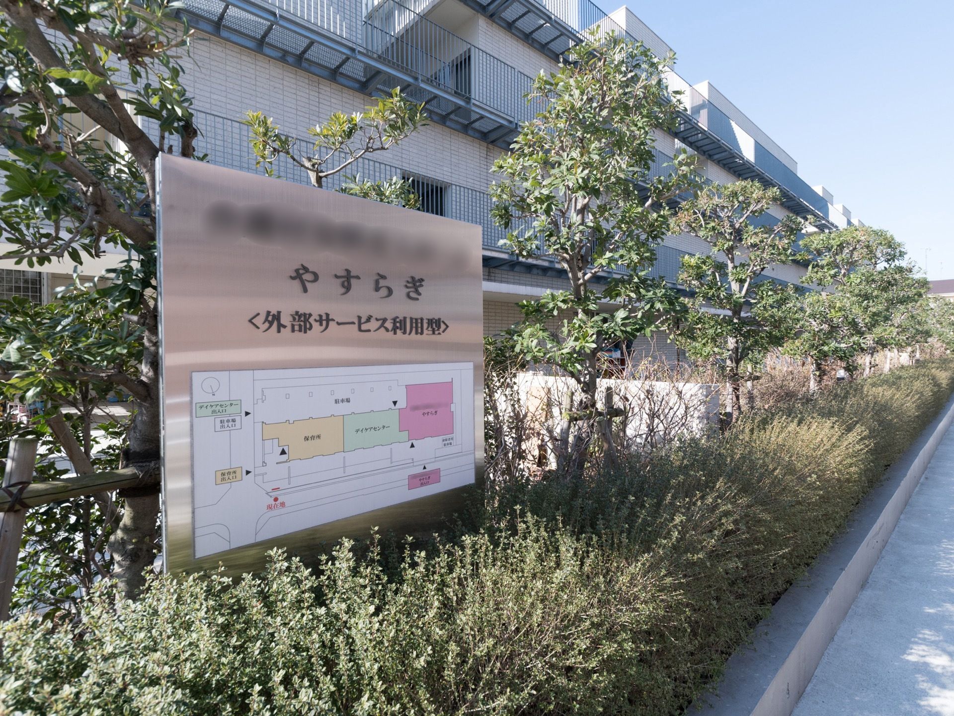 建物の前には植え込みがあり、その中に施設の看板が立っている。施設名称のほか、全体の見取り図も付いている。