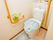 白い壁にオレンジの手すりが完備されたトイレ。便座カバーとトイレマットは、水色できれいである。緊急ブザーもすぐ手の届くところに設置されている。