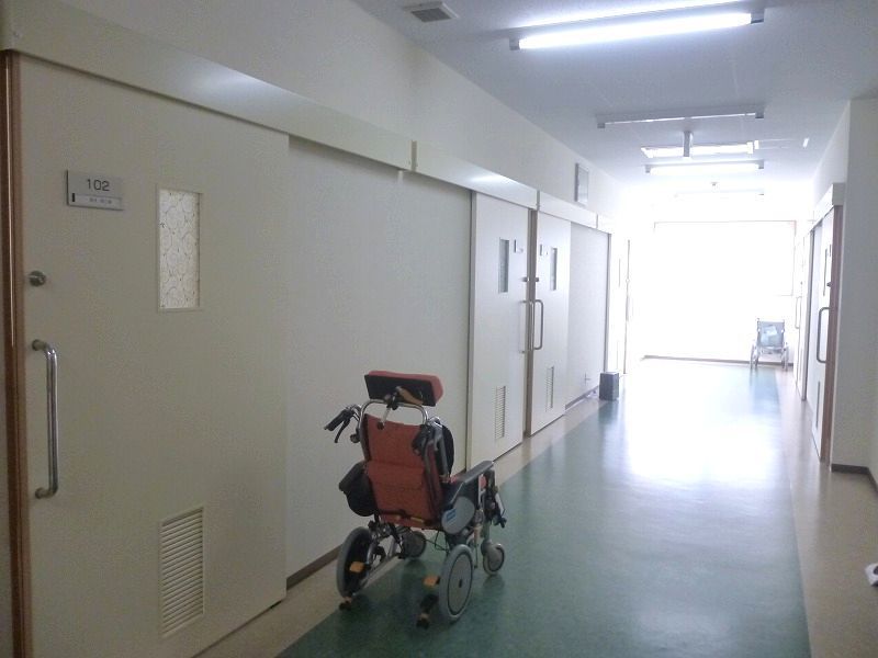 居室のスライドドアが並ぶ廊下は、幅が広く段差のない設計になっており、車椅子が置かれている。明るく白い壁に囲まれている。
