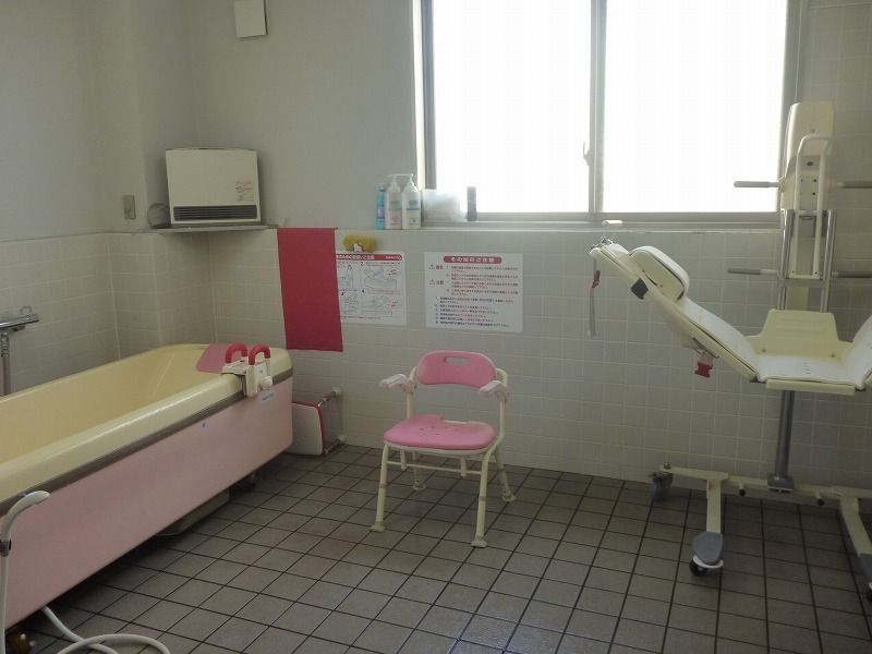 寝たままご入浴いただけるリフトと浴槽が設置された広い浴室。床と壁の下部はタイル張りで窓があり、椅子が置かれている。