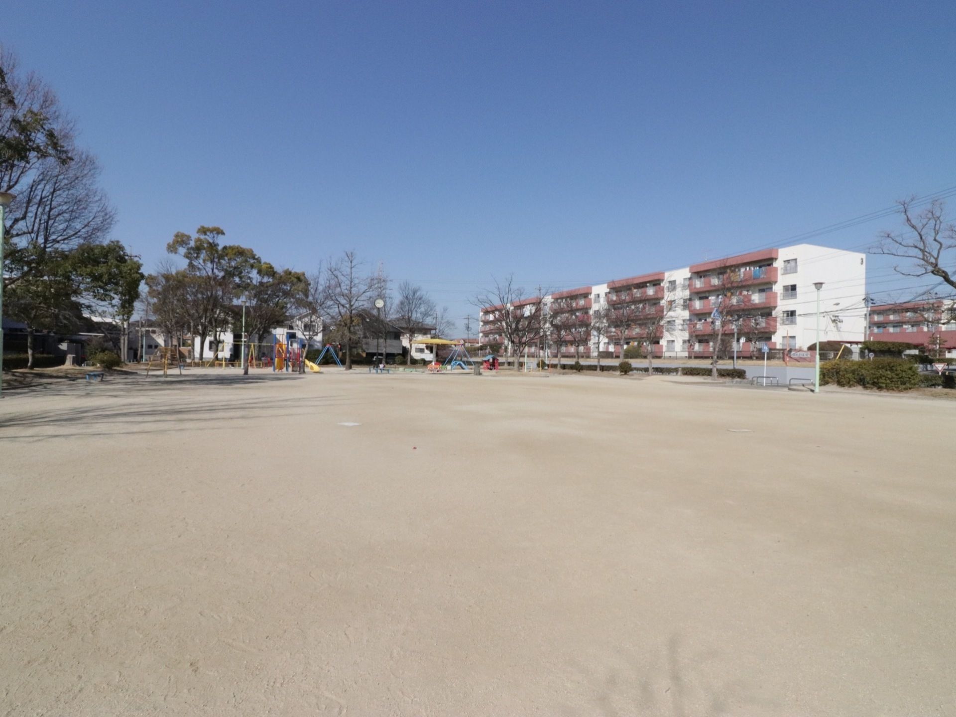 広い公園である。手前はグランウドになっており、子供の遊具が奥の方に設置されている。青空が広がり、右奥前方には６階建ての団地が見える。