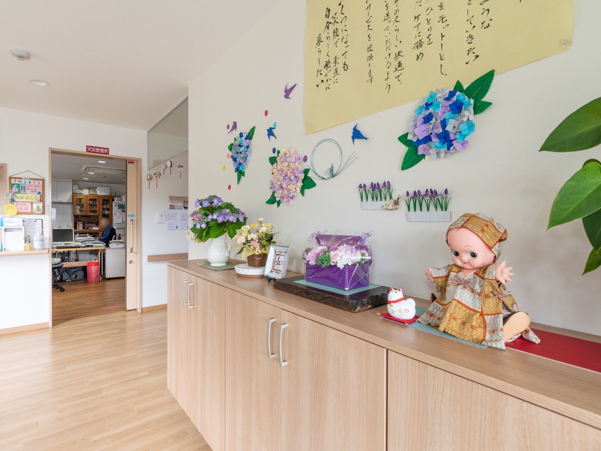 腰高ほどの収納がある。その上にはさまざまな花が花瓶に入って飾られている。壁には折り紙で作られた紫陽花や燕が貼ってある。