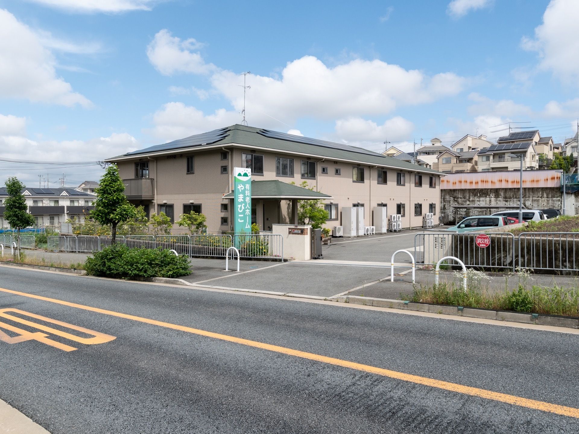 ２階建ての建物が見える。施設名が書かれた大きな看板が設置されている。施設前方は駐車場になっており、車が数台駐車している。