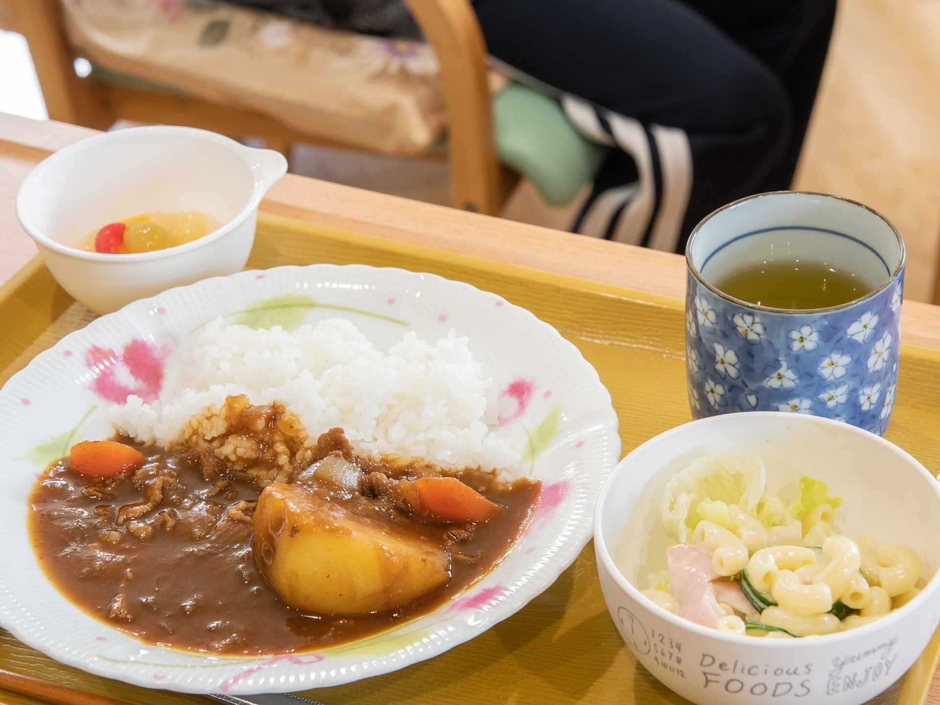 お盆の上に食事が用意されている。メニューはカレーライスとマカロニサラダ、デザートである。入居者様の足元が写っている。