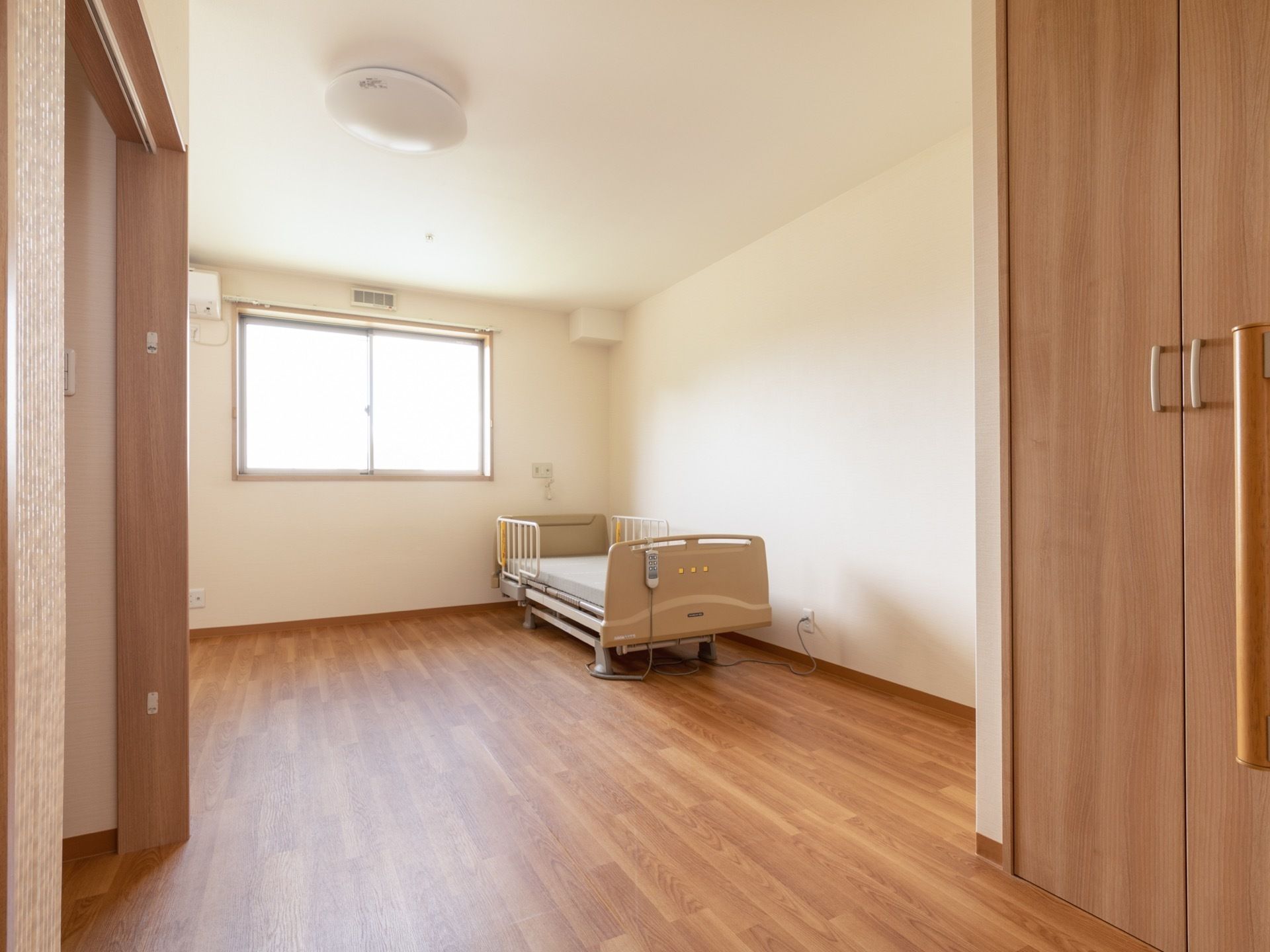 広々としたフローリングの部屋に電動ベッドが置かれている。腰高の窓があり、エアコンやシーリングライトなどが設置してある。