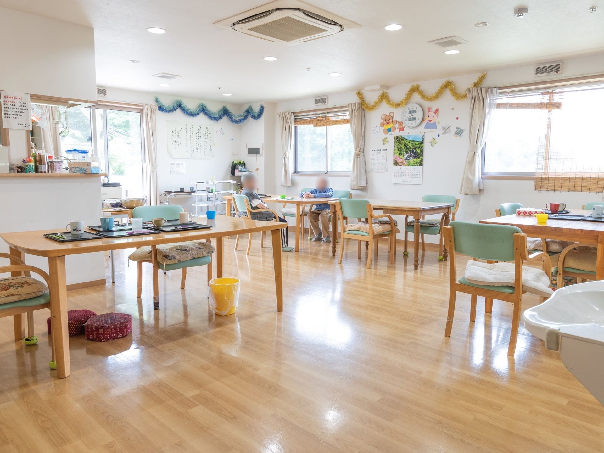 木目調のダイニングテーブルがあり、シートクッションが置かれた椅子が用意されている。窓にはカーテンやすだれが掛けられている。