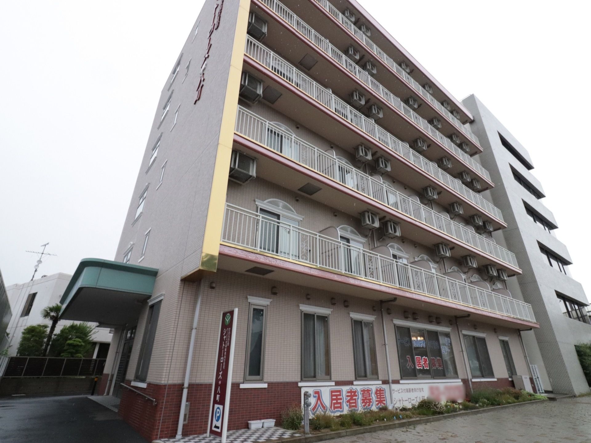 ピンク色を基調とした外壁で、窓枠のデザインに欧風の雰囲気が漂うモダンな建物である。玄関前はフラットで、壁に手すりが設置されている。