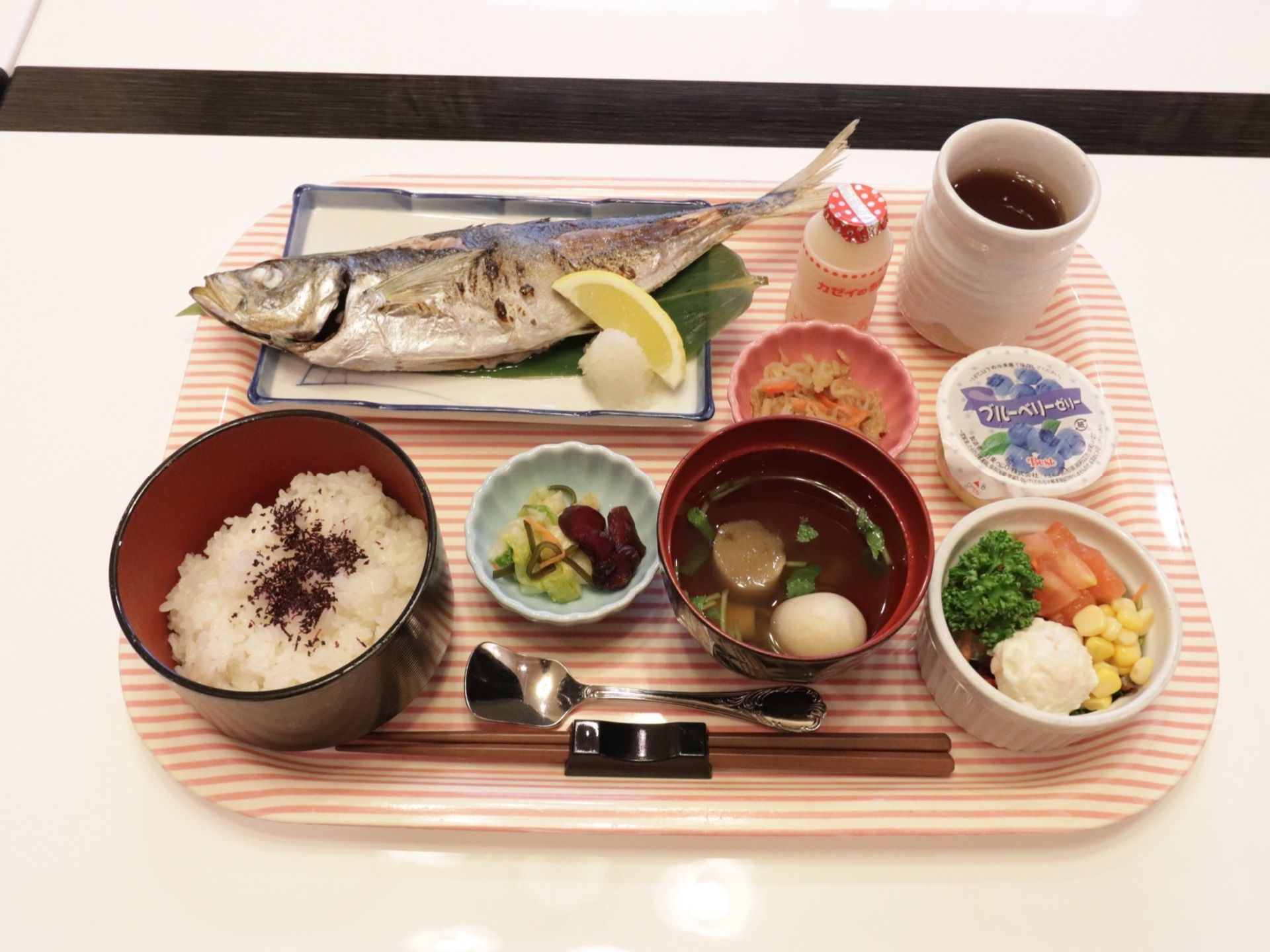 ボーダーのピンクのトレイに1人分の食事が並んでいる。ご飯、お吸い物、漬け物、サラダ、煮物の小鉢、焼き魚、デザートが用意されている。