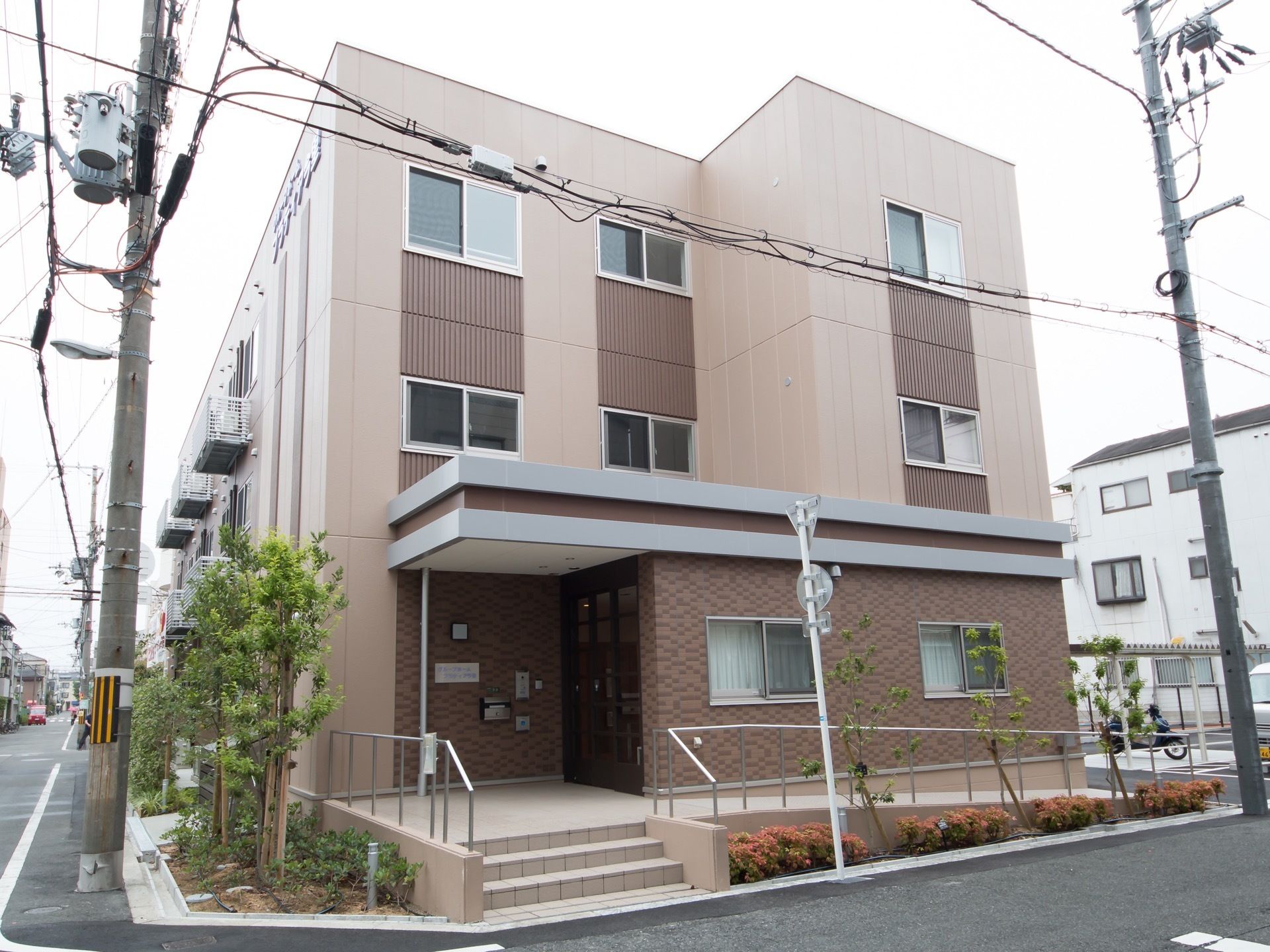 ３階建ての建物の入口側の画像。入口に繋がるスロープと階段があり、それぞれに手すりが取り付けられている。