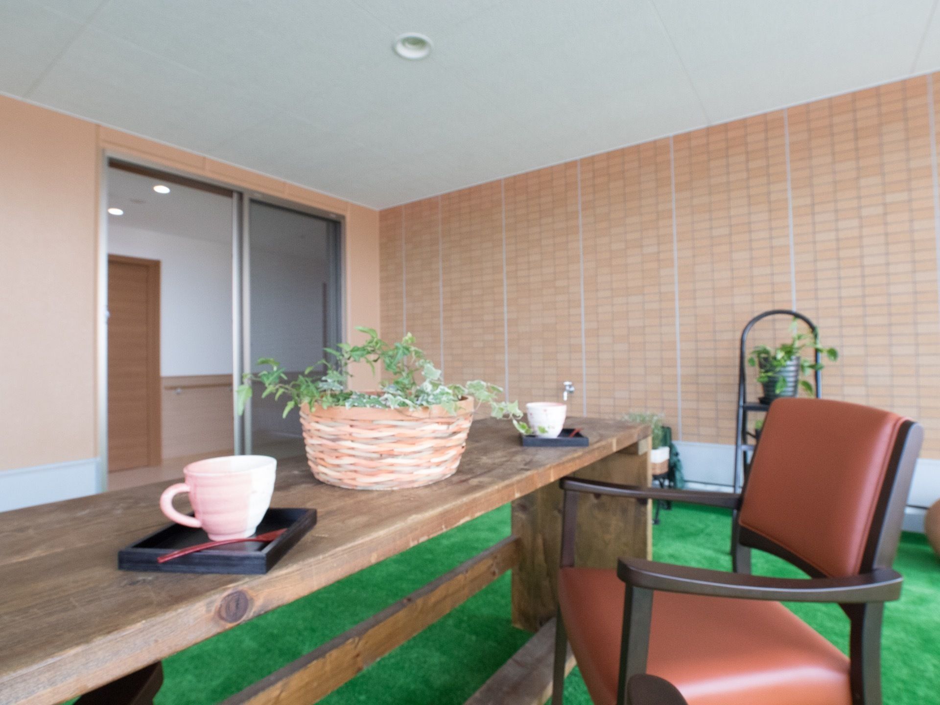 人工芝が敷かれ、テーブルと椅子が置かれている。観葉植物などが飾られている。テーブルの上にカップが2つある。