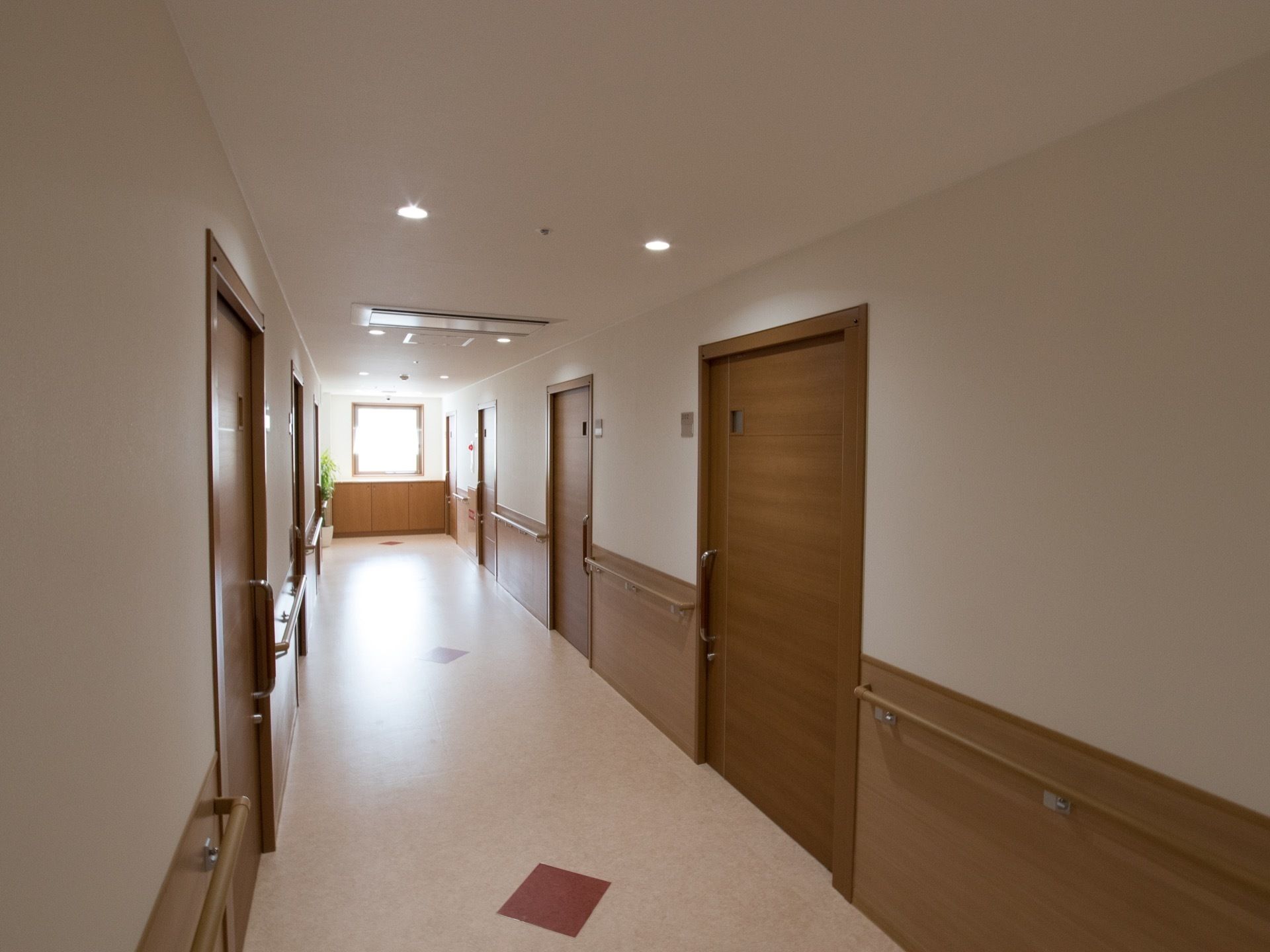 通路の両端に手すりが設置されている。居室ドアは引き戸式である。通路奥に窓があり、陽射しが入ってきている。