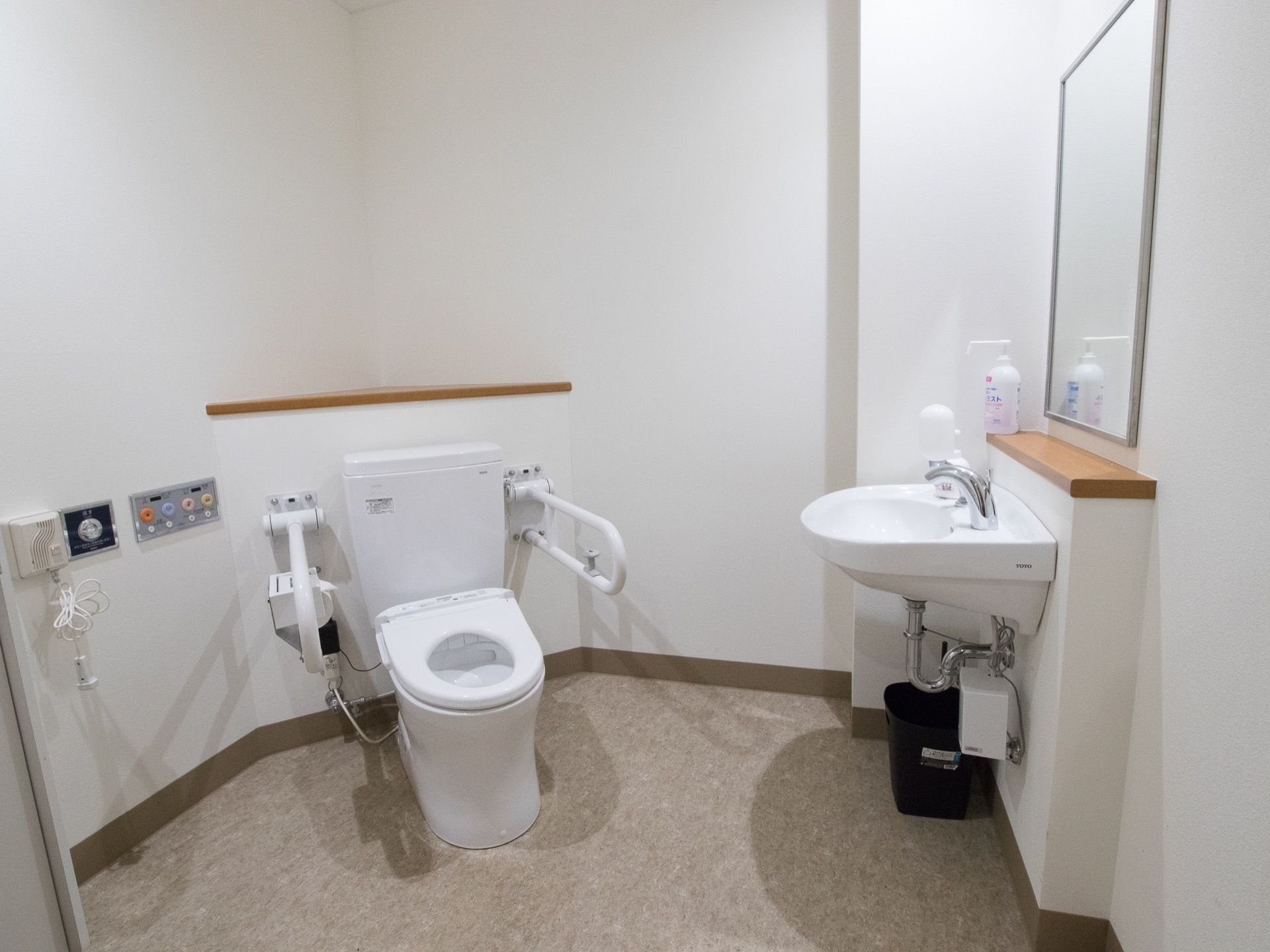 トイレの隣に脚なし洗面台がある。棚の上に手指用消毒スプレーが置いてある。床にゴミ箱が置かれている。壁に鏡がある。