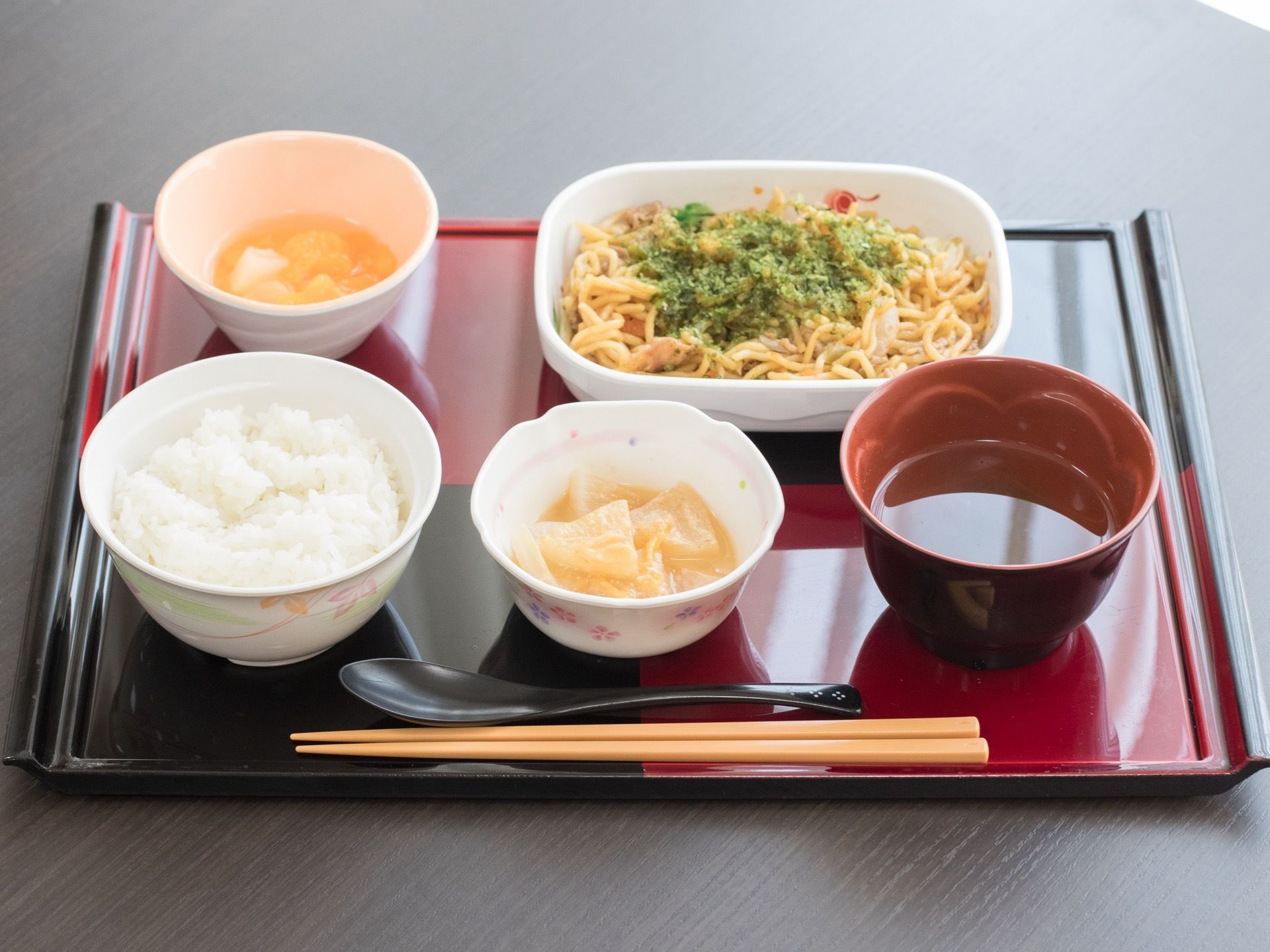 トレイの上にご飯、汁物、焼きそば、小鉢、フルーツ、箸、スプーンが置かれている。白地に花柄などのついた食器を使用している。