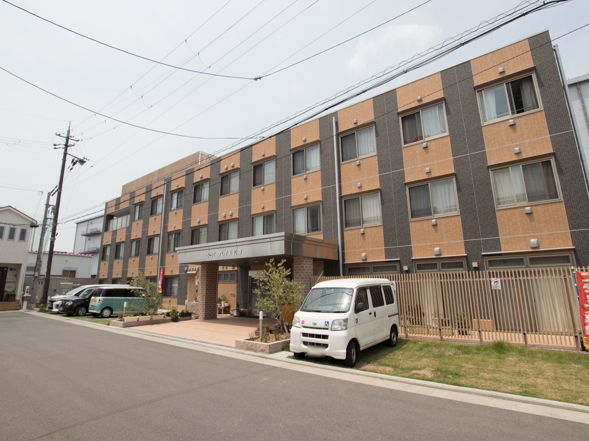 建物の前に縦格子の柵が取り付けられている。玄関を挟んで両側に芝生の駐車スペースがあり、車が数台停まっている。