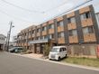 建物の前に縦格子の柵が取り付けられている。玄関を挟んで両側に芝生の駐車スペースがあり、車が数台停まっている。