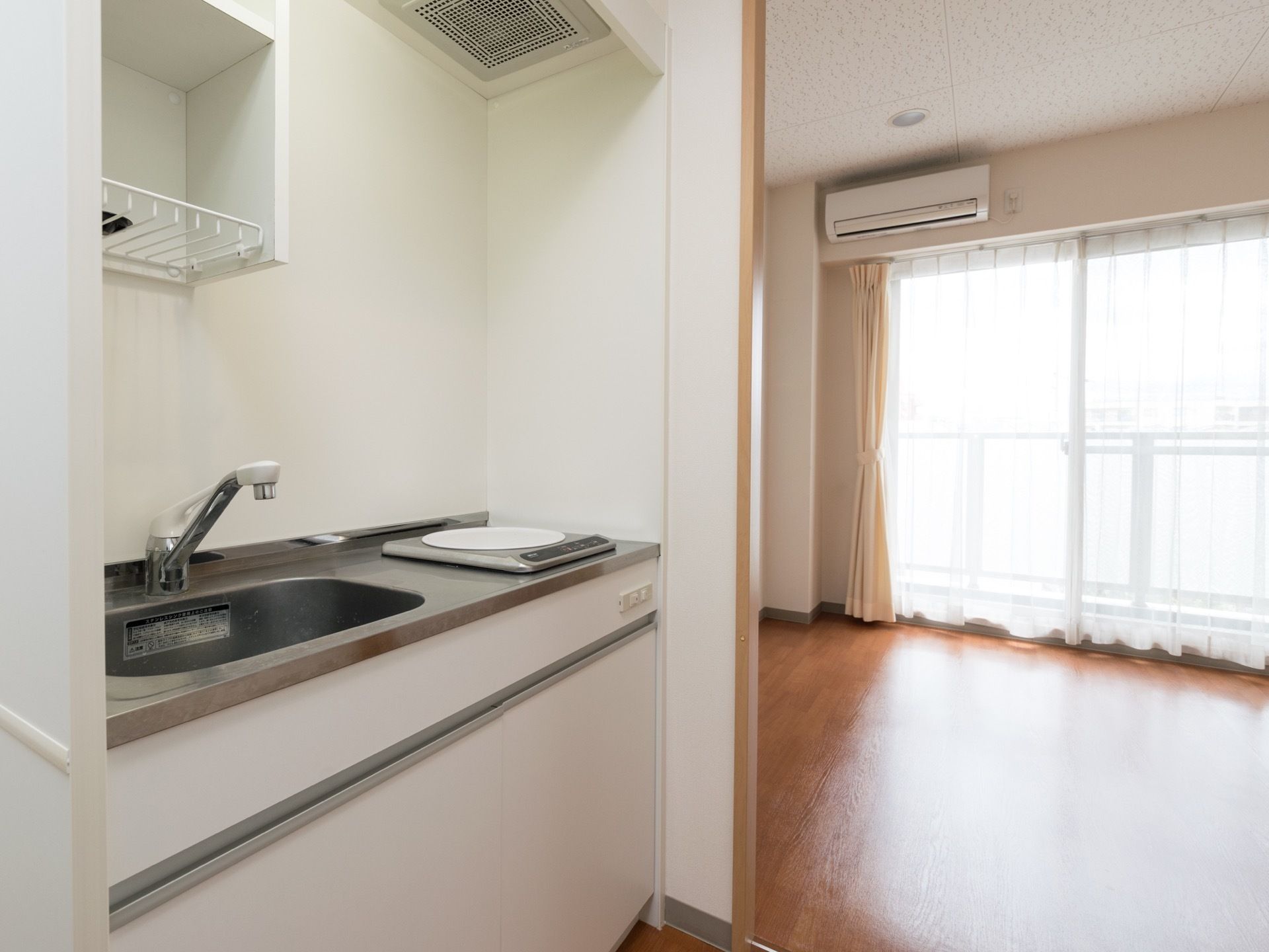 手前には、コンロが一基取り付けられたキッチンシンクが見える。奥には、日当たりのよさそうな、フローリングの部屋がある。