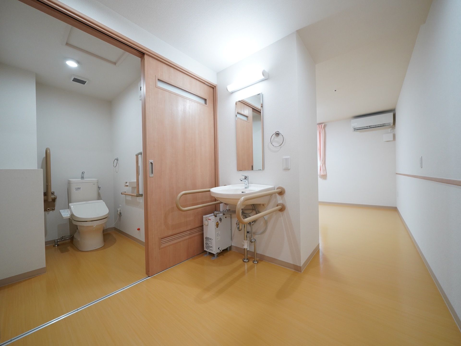 居室に設置された温水洗浄便座付きの広いトイレである。ドアは引き戸式となっている。入口付近に洗面台が置かれている。