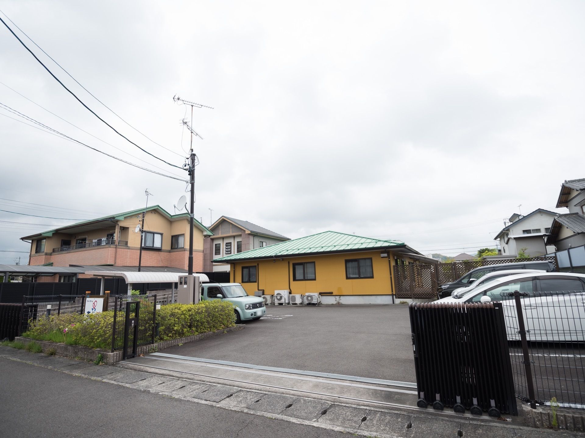 黄色の壁にグリーン調の屋根、ブラウンのサッシでアクセントを入れた平屋建ての施設建物である。施設手前には、駐車場も併設されている。
