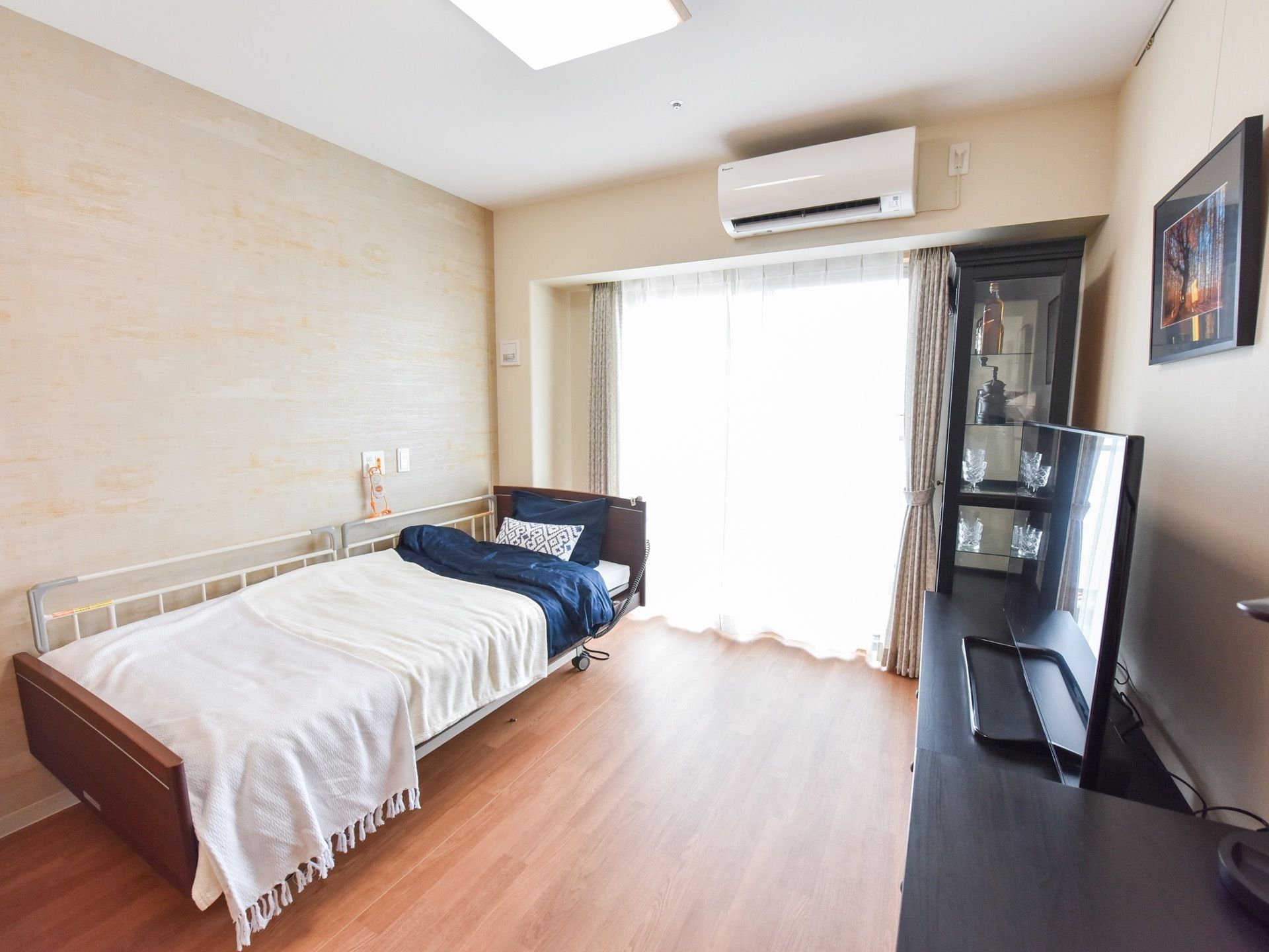 白い壁紙とフローリングの部屋に、ベッドやテレビ、ガラス張りの戸棚などが置かれている。窓際にはエアコンが設置されている。