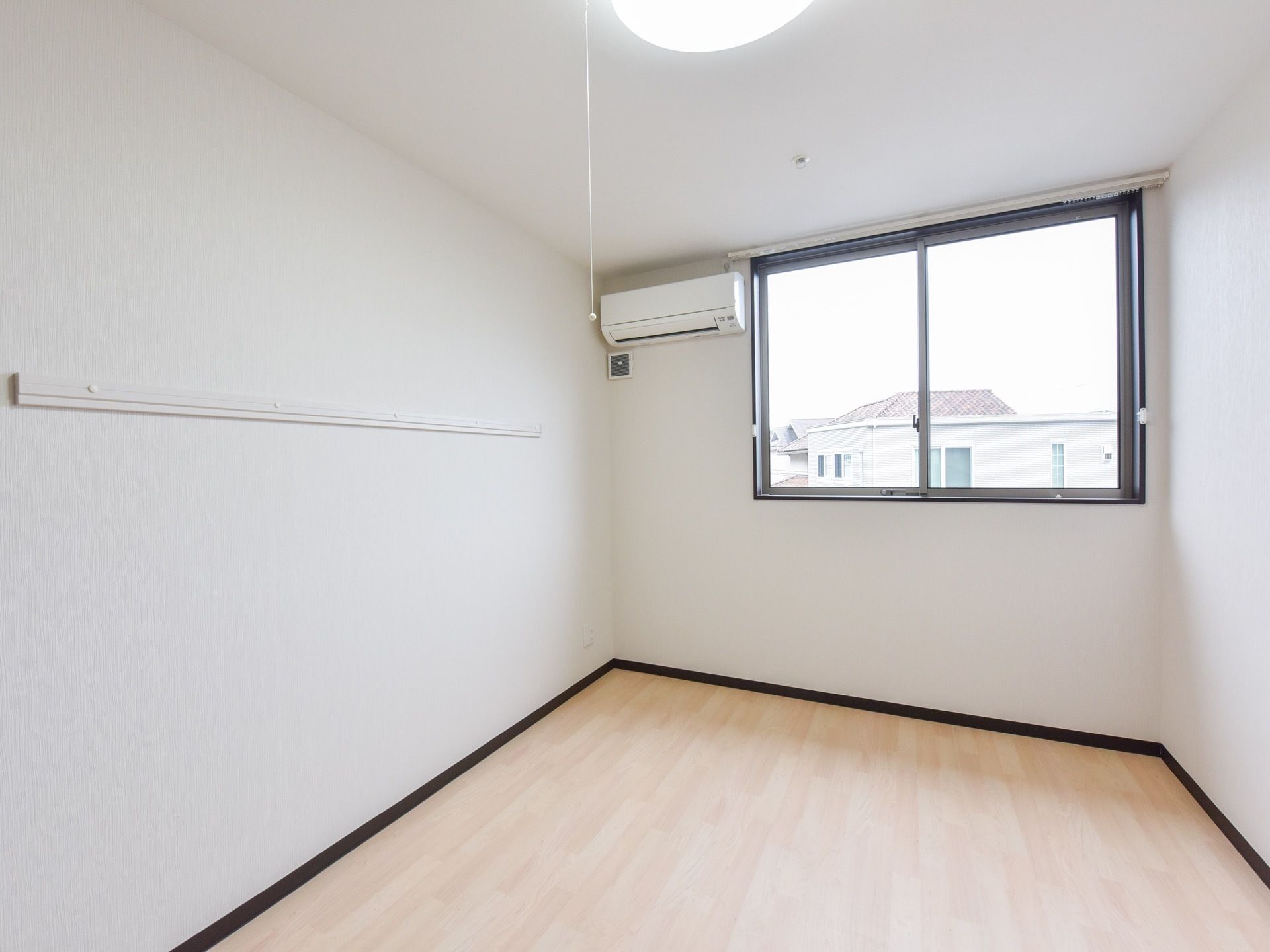 明るい色のフローリングの床と白色の壁の、すっきりとした室内である。窓の左上にエアコンが設置されている。