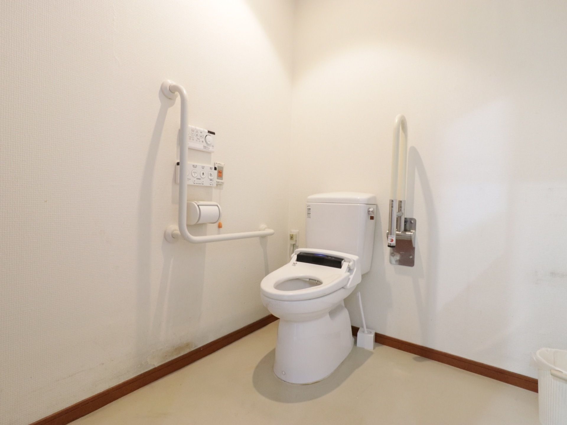 隅に温水洗浄便座が置かれている。白い壁に、固定式と可動式の手すりが取り付けられている。広いトイレである。