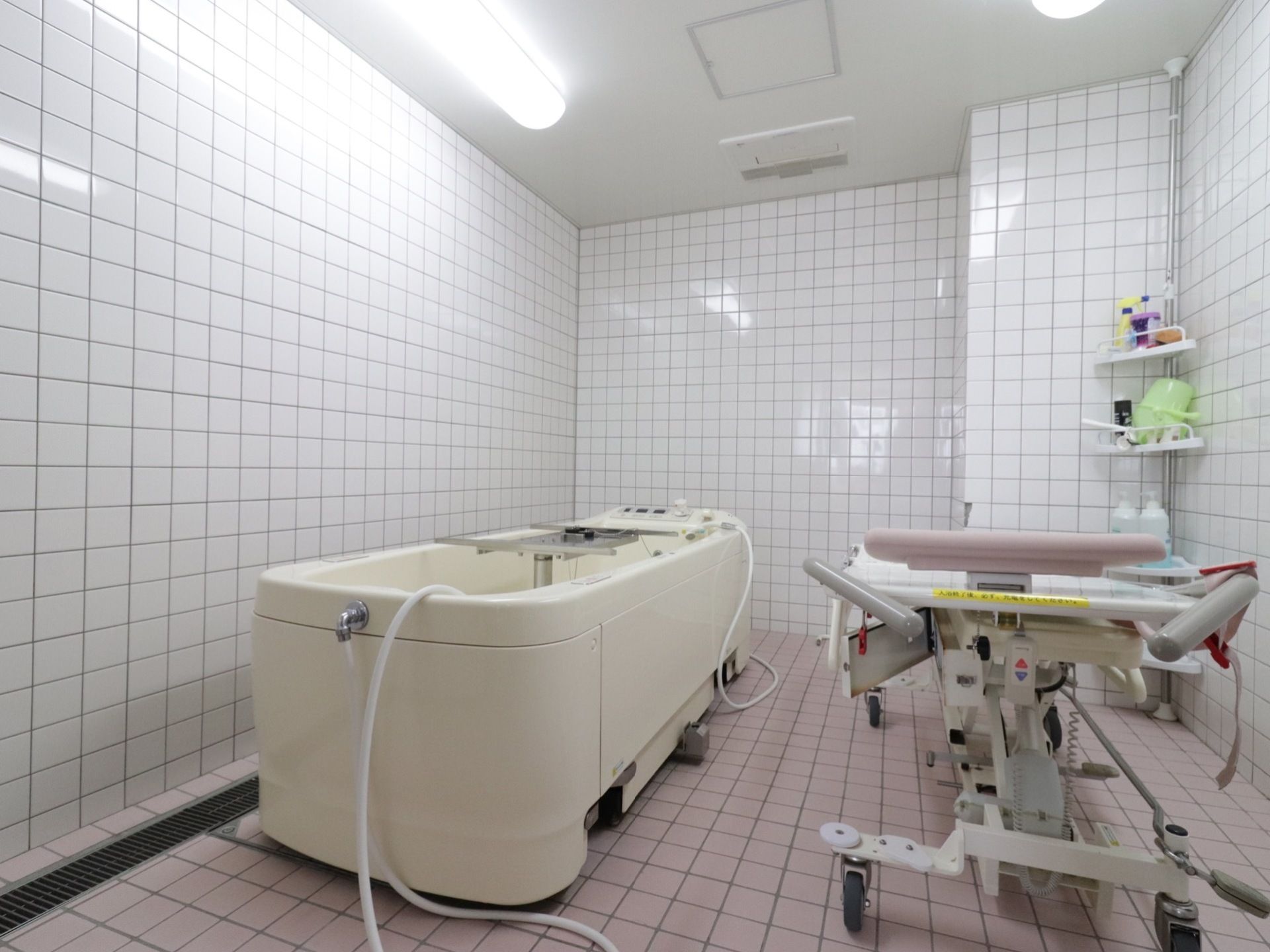淡いピンクの床に白の壁の全面タイル貼りである。介護入浴機器が置かれている。明るい照明のある浴室である。