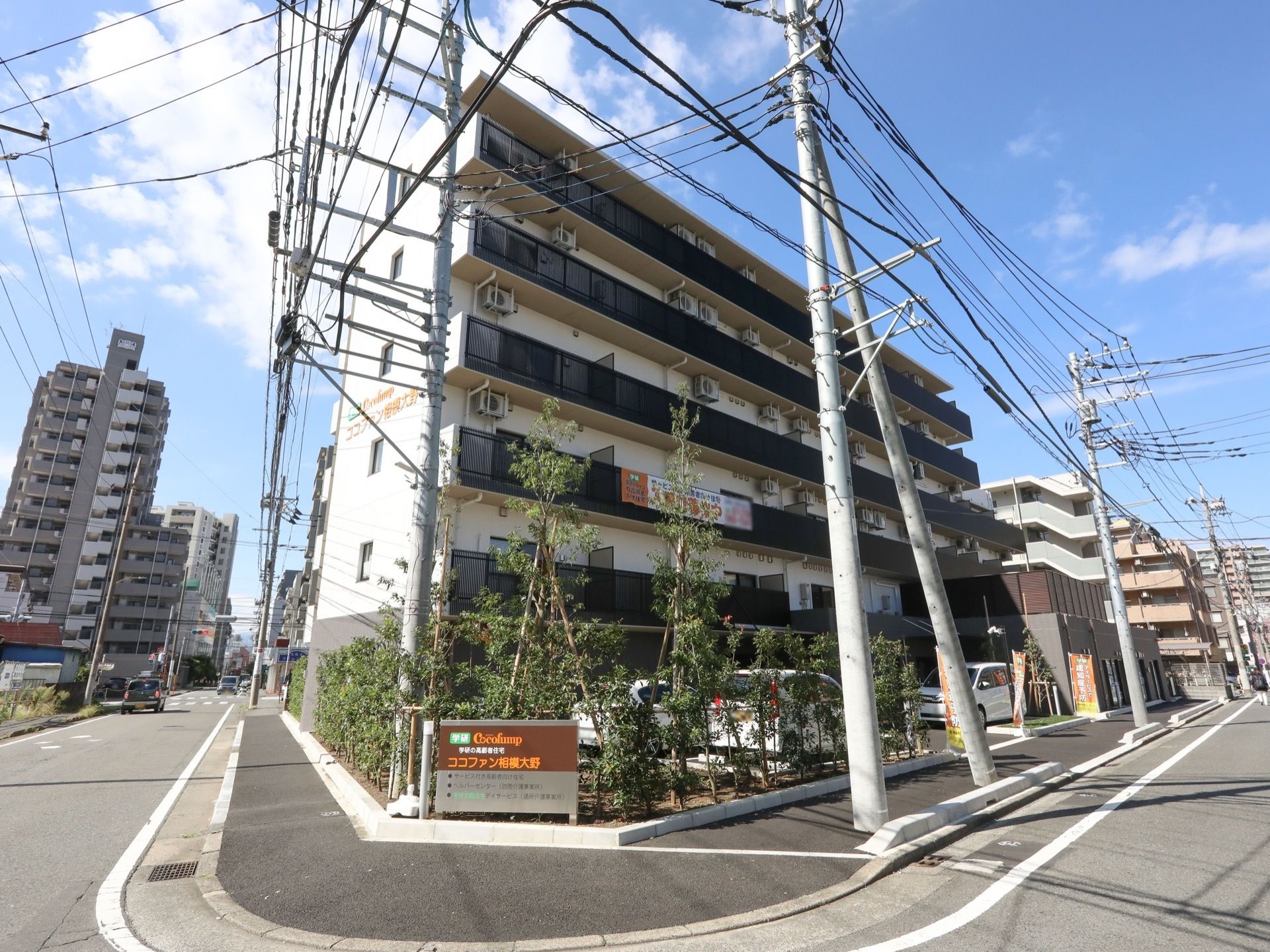 道路の曲がり角に面した場所に、施設の案内板がある。建物の前には建物の２階以上の高さのある植物が植えられている。