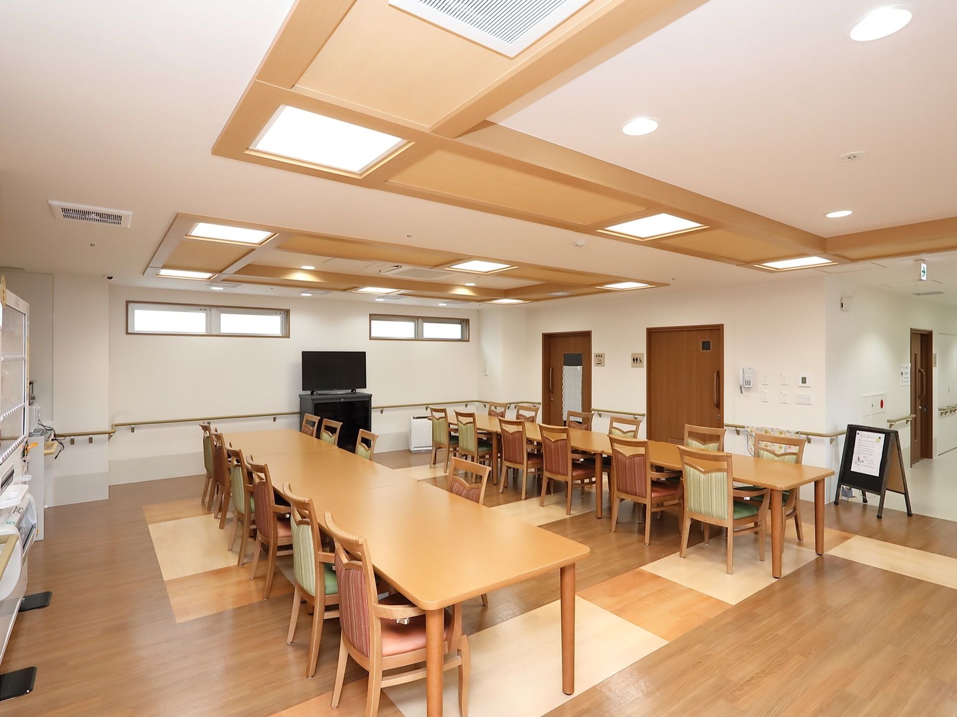 大きな照明器具が天井に複数箇所埋め込まれていて、明るい食堂である。角が丸くなっている長テーブルと、赤や緑の布が施された椅子が並べられている。