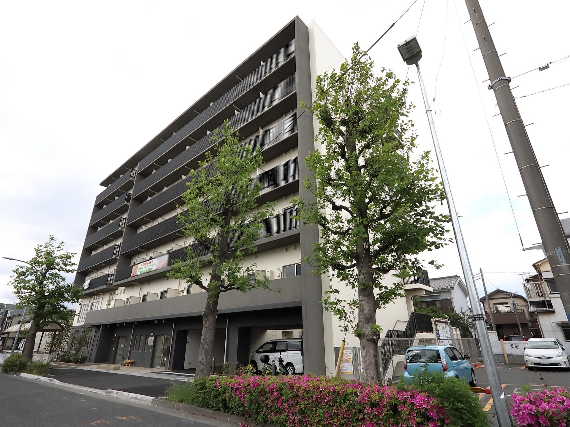 道路に面してそびえたつ７階建てのビル。建物の１階部分にガレージがあり、右隣にはコインパーキングがある。