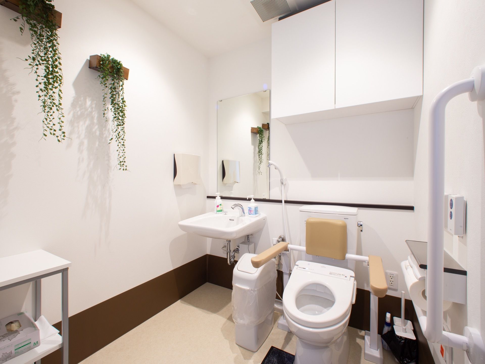 白を基調とした広い共用トイレである。洗面台や手すり、背もたれ付きアームレストが完備されている。ゴミ箱が置かれている。