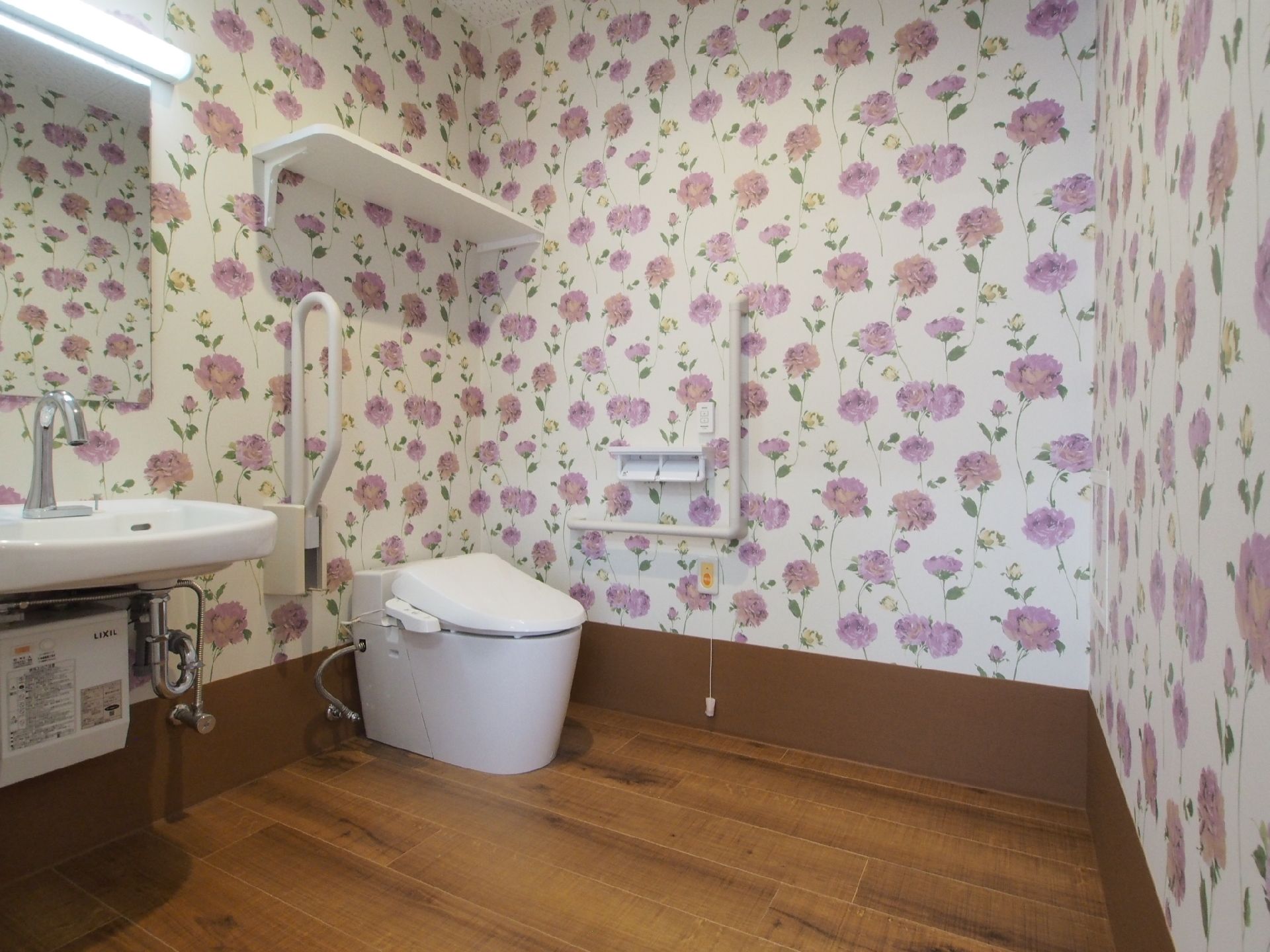 トイレは広く手洗い場も付き、壁一面に花柄の壁紙が貼られ、手すりも設置されている。