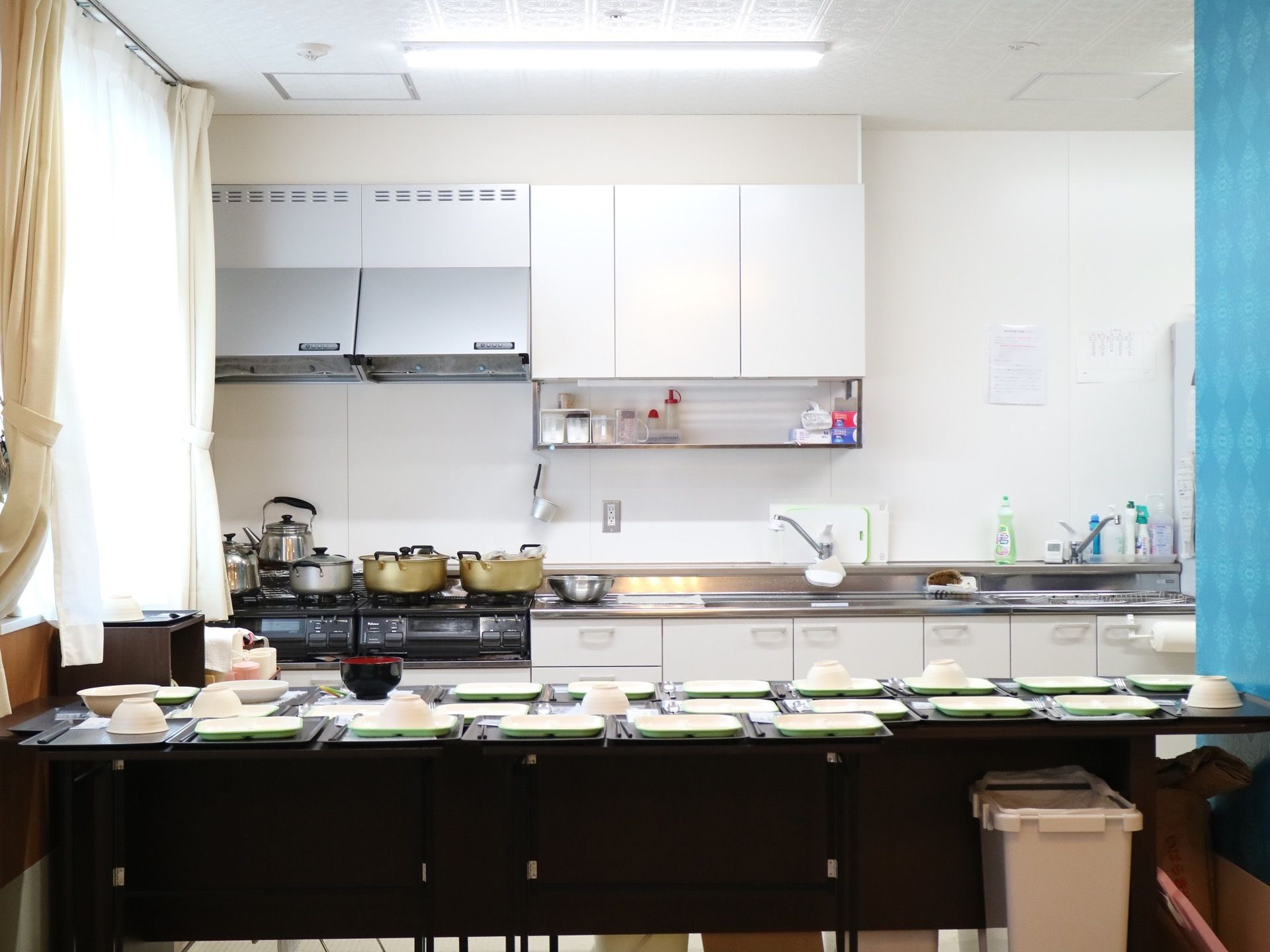 奥にガスコンロやシンクなどが備え付けられたキッチンがある。その手前にはテーブルがあり、テーブル上にたくさんのトレイと食器が並べられている。
