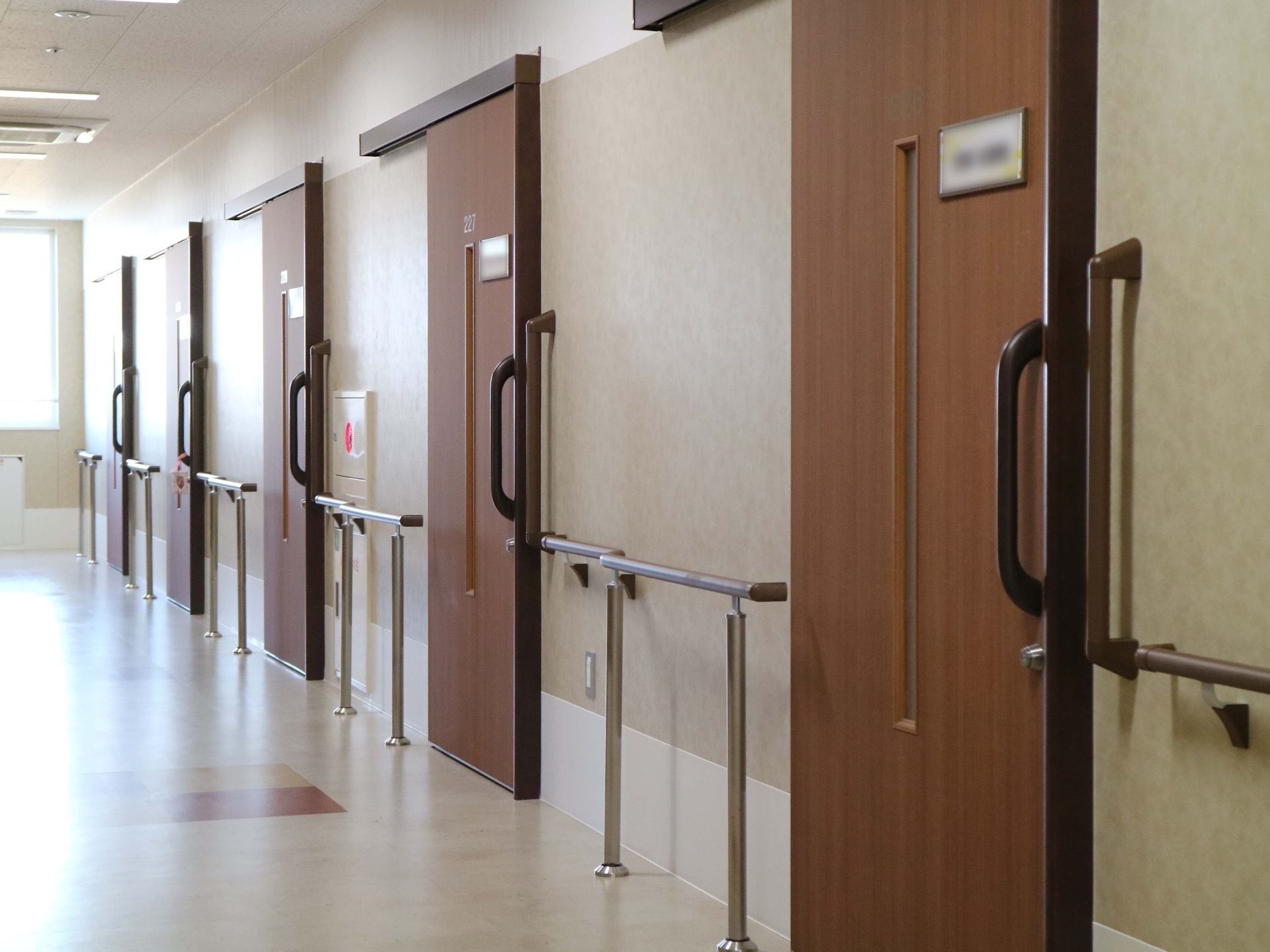 引き戸の扉がたくさん並ぶ通路。扉の周囲の壁には手すりがある。通路の奥には窓があって、天井には空調の吹き出し口があるのが見える。