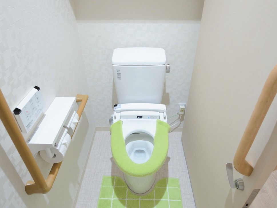タンク付きの洋式便器が設置され、黄緑色の便座カバーが掛けられている。壁に手すりと２連タイプのペーパーホルダーが取り付けられている。