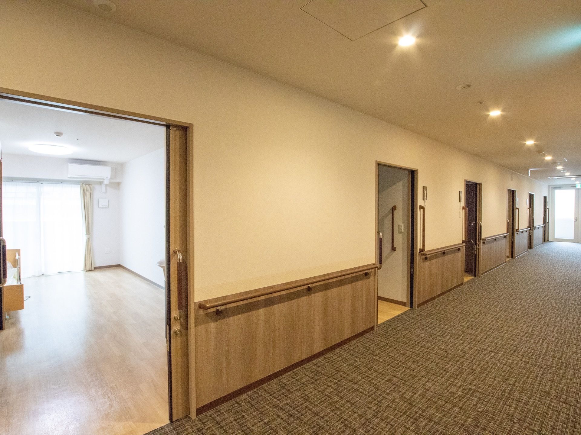 入居者様の居室が並ぶ廊下。床は絨毯になっており、壁紙と同系色で統一されている。壁には手すりが設置されている。