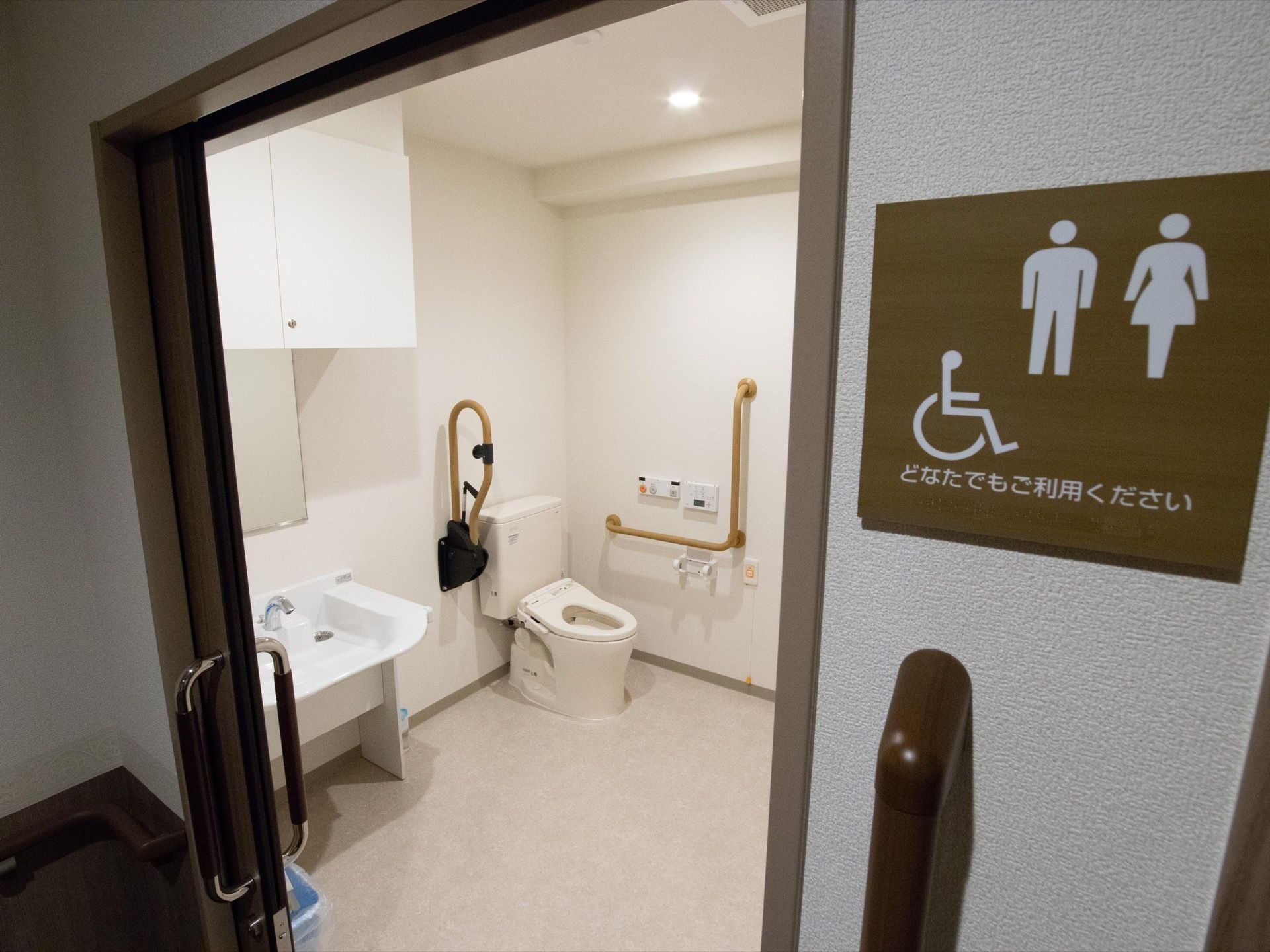 ダークブラウンの枠ぶちのついた広いトイレがある。トイレの標識も同系色で揃えてある。洗面台には鏡と棚があり、手すりが設置されている。