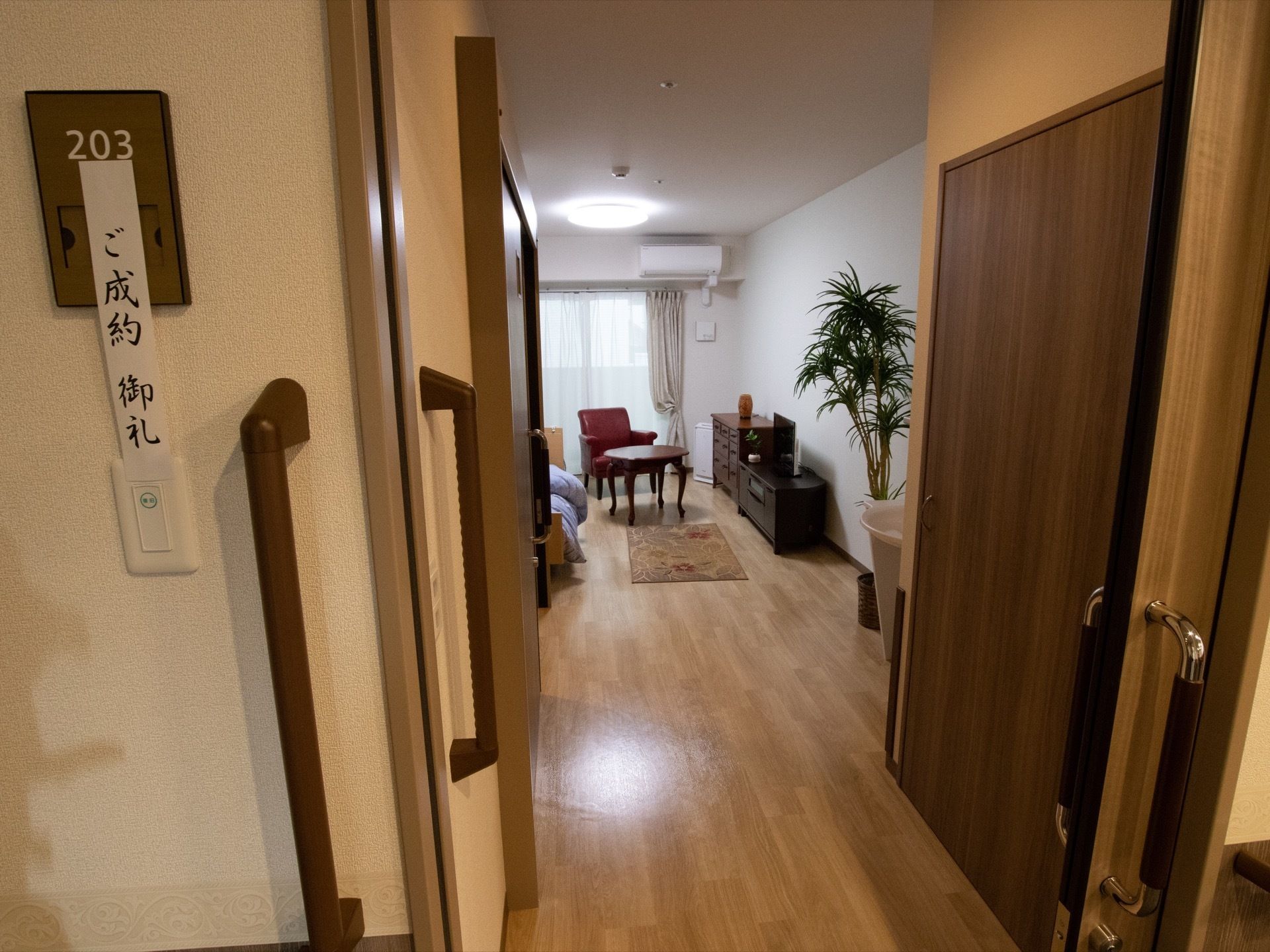 スライド式の扉の先に、入居者様の居室がある。ソファ、テーブル、チェスト、テレビなどの家具が見える。観葉植物が置かれている。