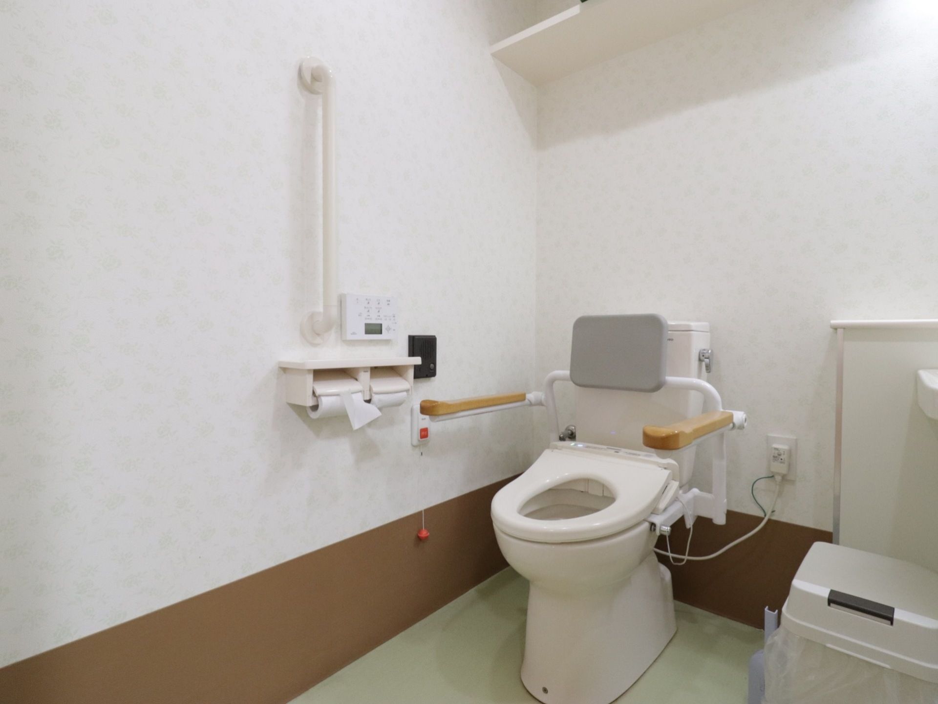トイレは白で統一されていて清潔感がある。便座には蓋がなく背もたれが付いている。左右に肘置きも設置されている。