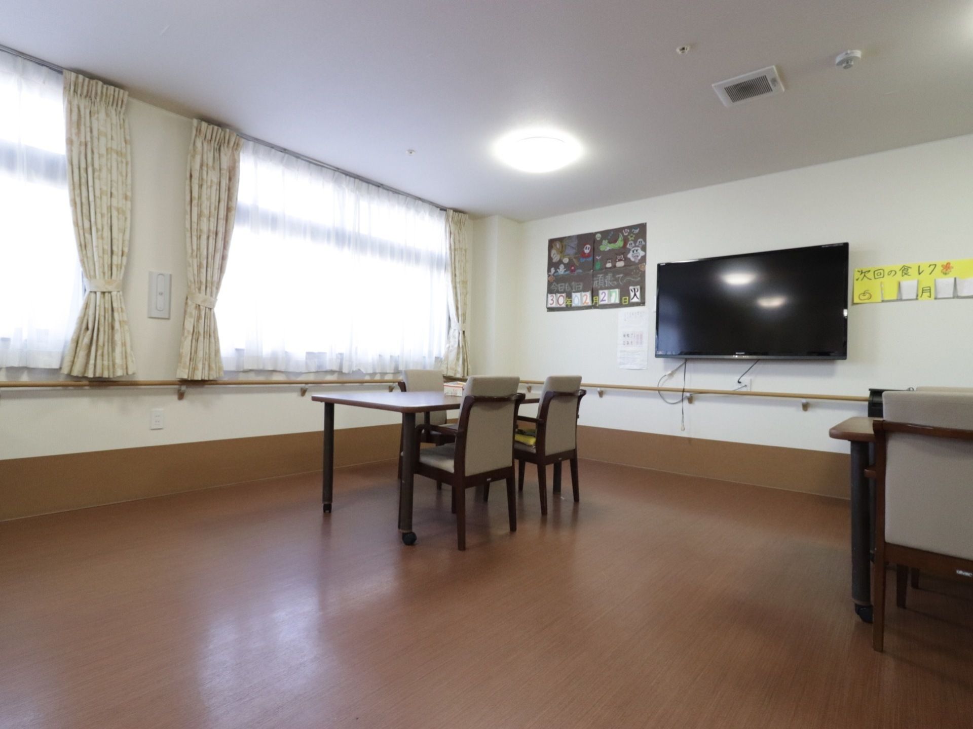 フローリングの広いスペースにダイニングテーブルがある。壁には大型のテレビや手作りのカレンダーがある。