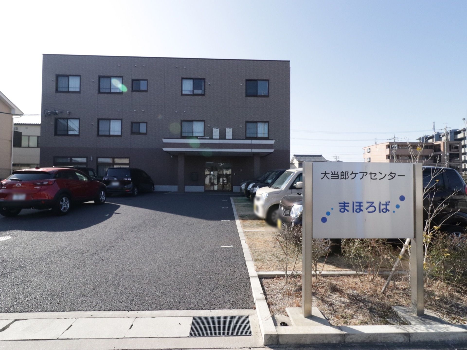 3階建ての施設の外観はシンプルになっている。他建物の前には駐車場があり数台の車が停められる。玄関前には屋根がある。